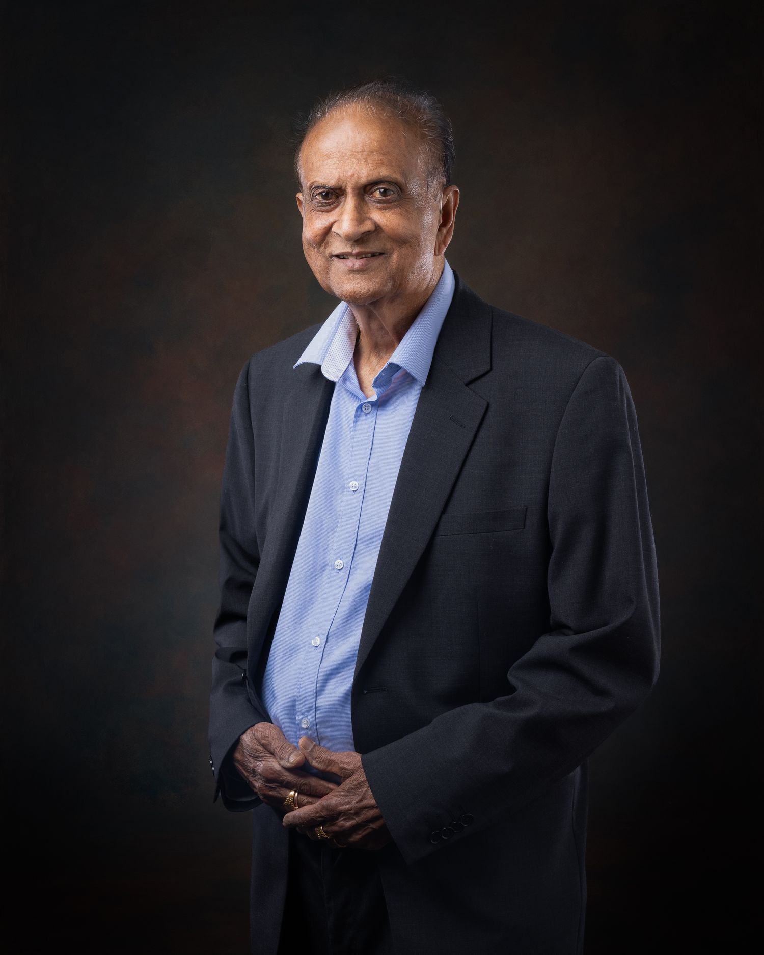 Professional studio portrait of a smiling person wearing a light blue dress shirt and dark blazer against a shadowy background.
