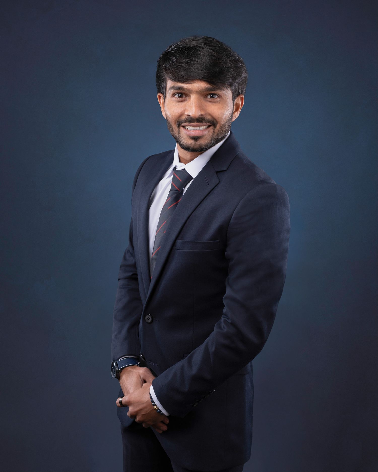 Professional studio portrait against dark blue background showing a well-dressed person in a navy suit and tie smiling.
