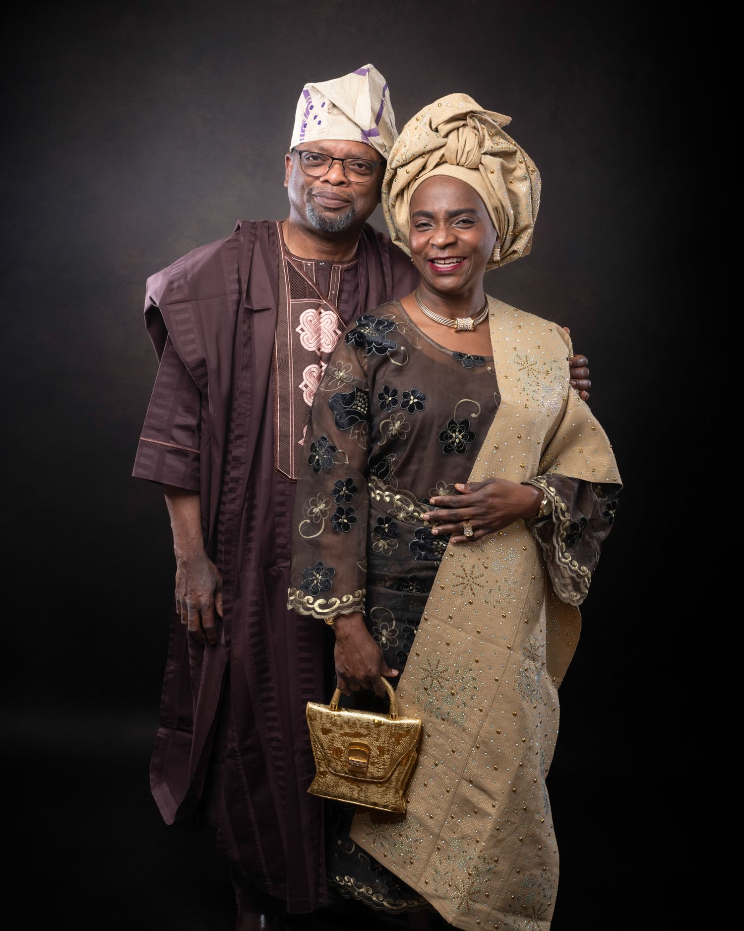 A couple in traditional Nigerian attire consisting of embroidered agbada and gele poses against a dark background.