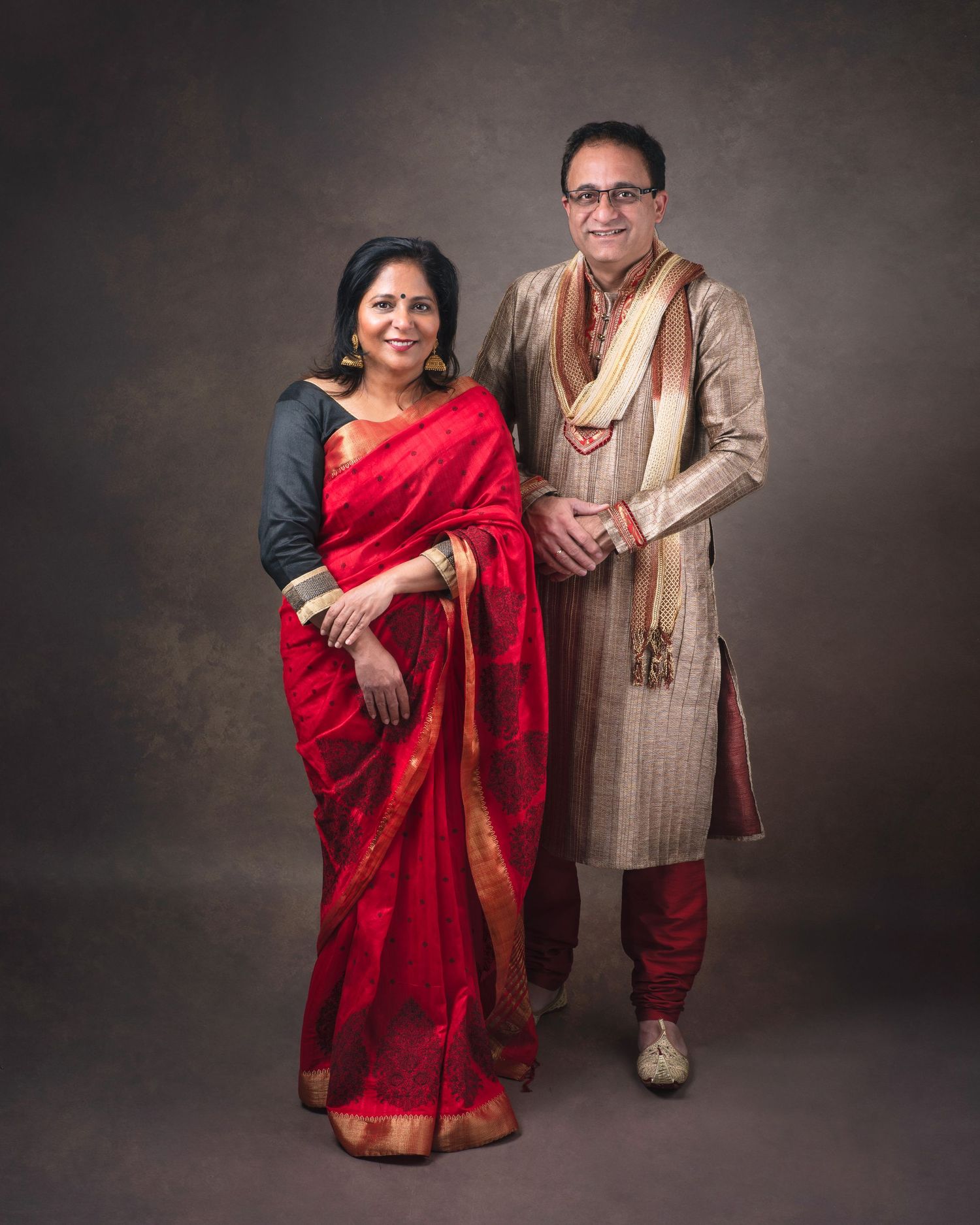 Couple in traditional Indian attire posing against dark backdrop wearing red sari and beige sherwani.