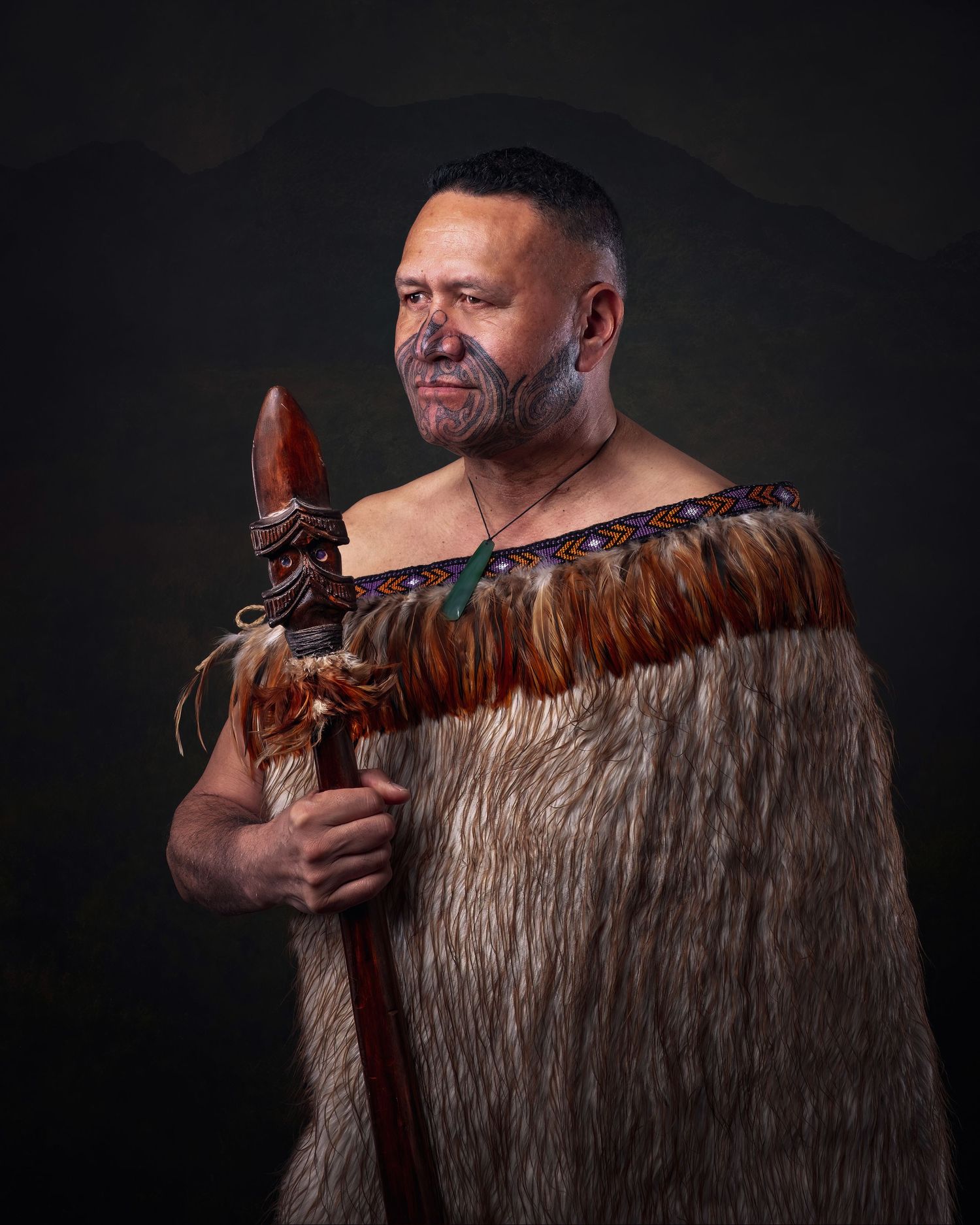 Traditional Maori warrior wearing ceremonial feather cloak against dark background in dramatic portrait photography.