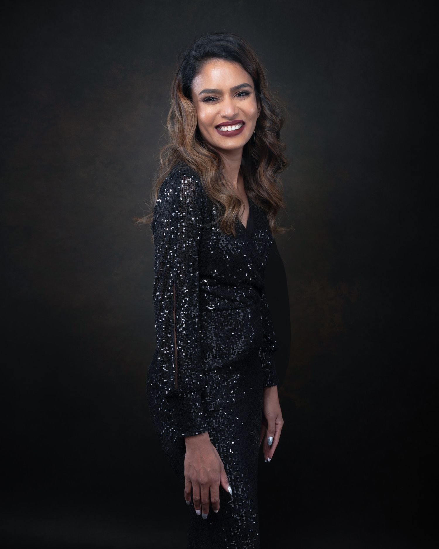 A figure in a sparkling black sequined dress poses against a dark background in a glamorous studio portrait.
