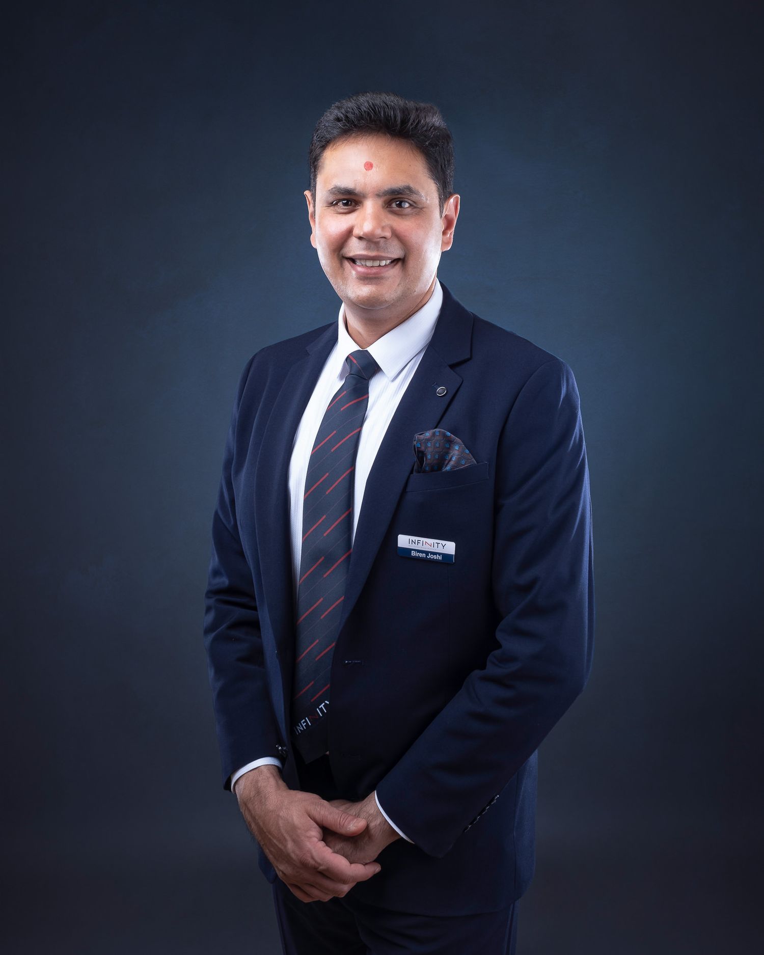 Professional corporate headshot of person in dark navy suit and tie against blue-gray backdrop.