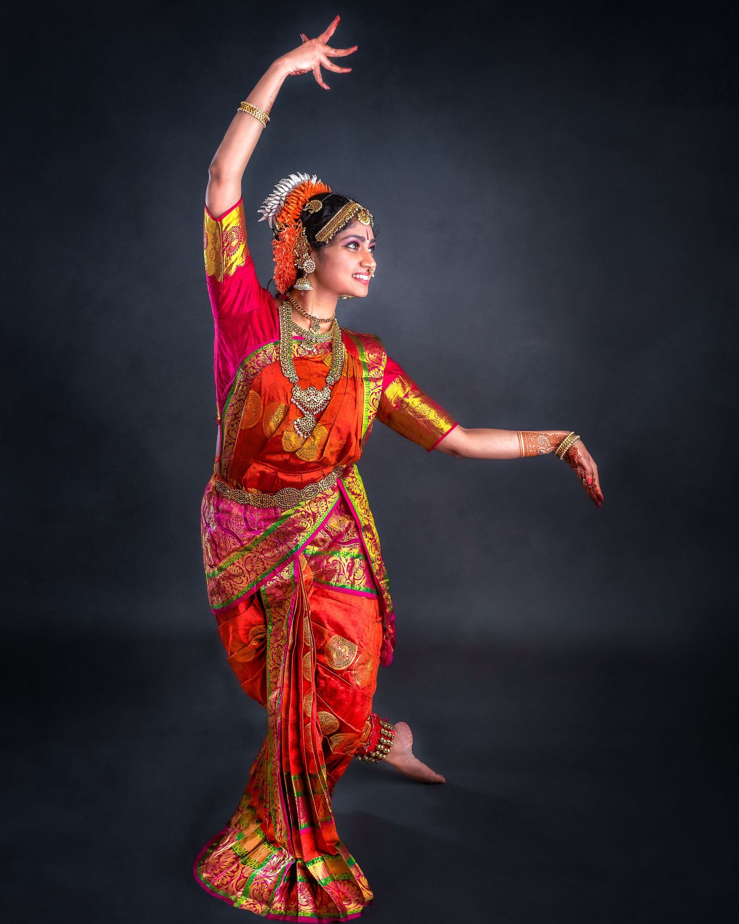 Classical Indian dancer in red and gold sari performs graceful pose against dark background with upraised arms.