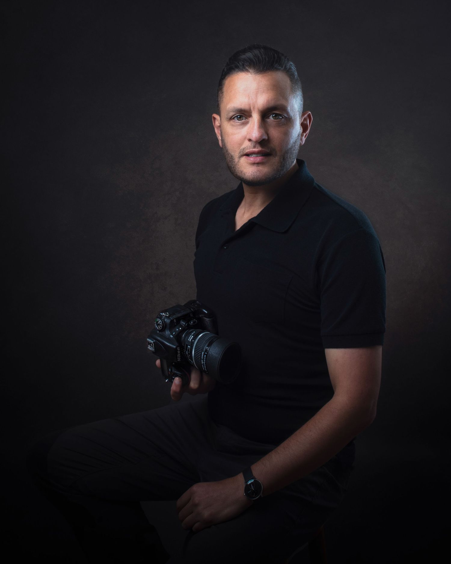 Professional portrait of a photographer wearing a black polo shirt holding a camera against a dark background.
