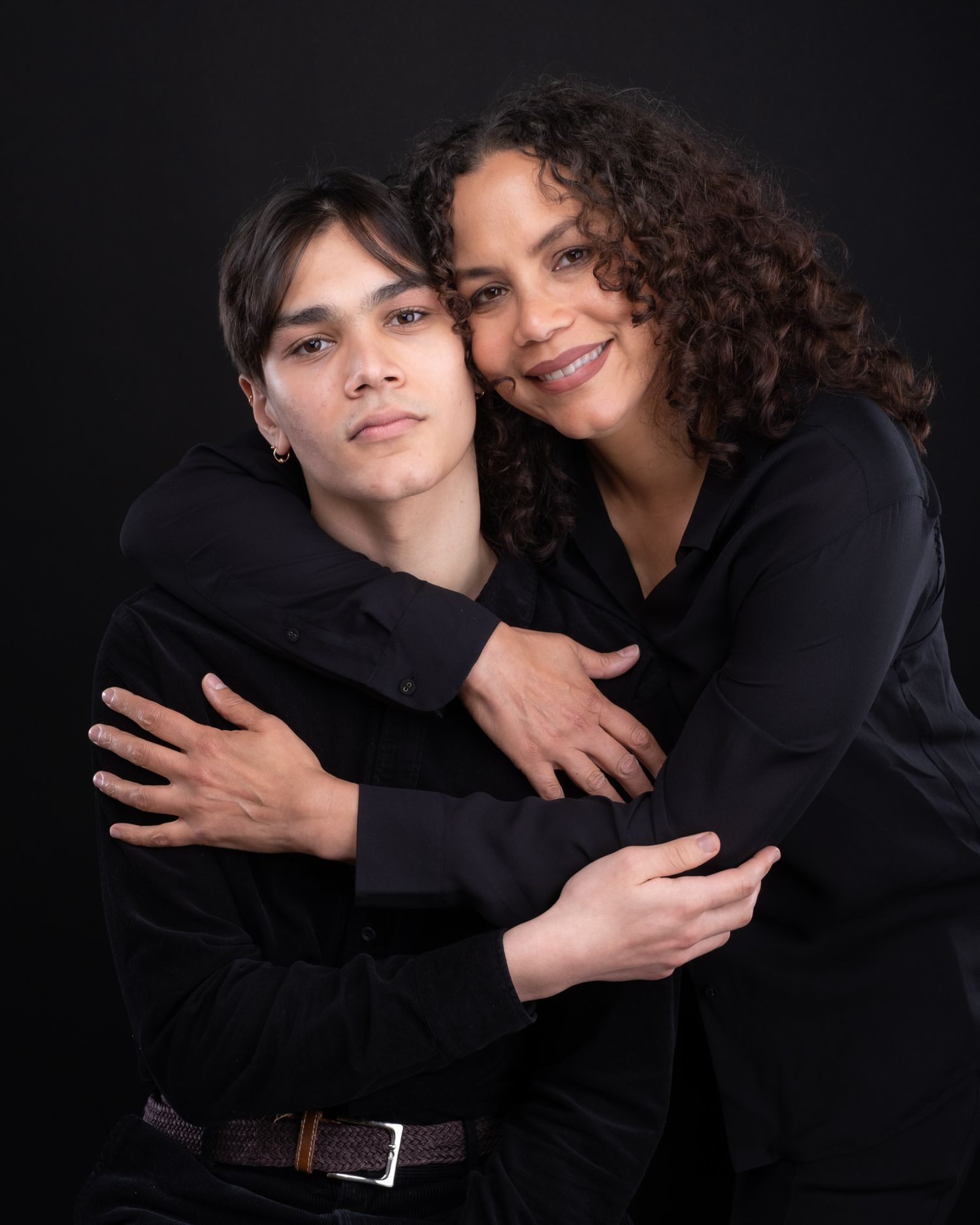 Studio portrait of two people embracing and smiling while wearing black clothing against dark background.