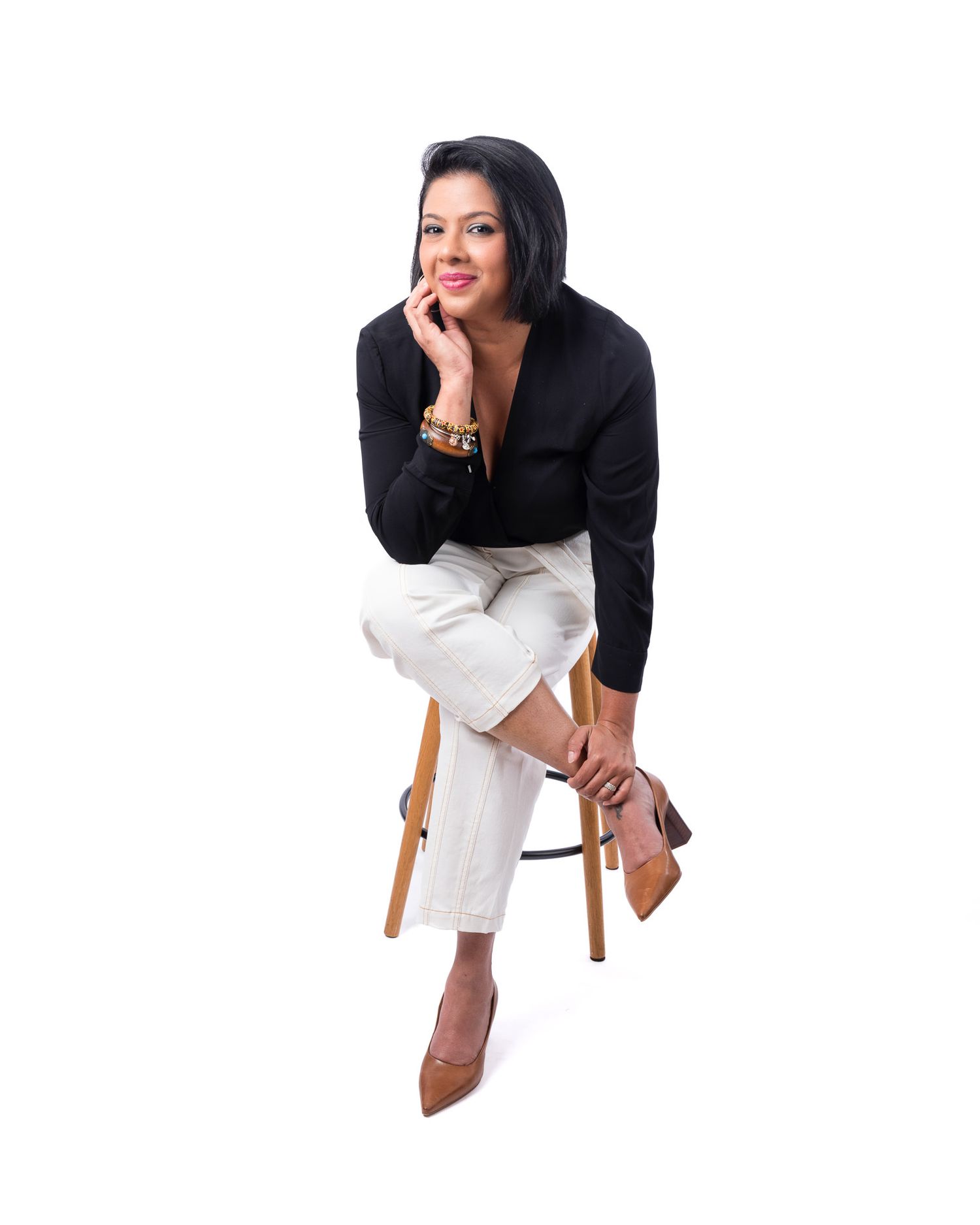 A woman in elegant business attire sits on a wooden stool wearing a black top, white pants and brown heels.