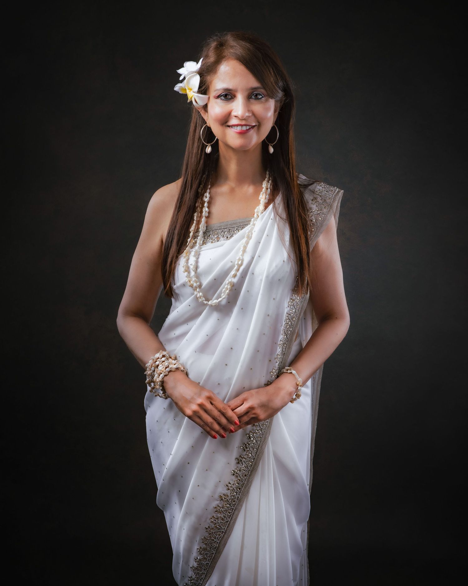 A person in an elegant white sari adorned with pearls and a flower poses gracefully against a dark background.