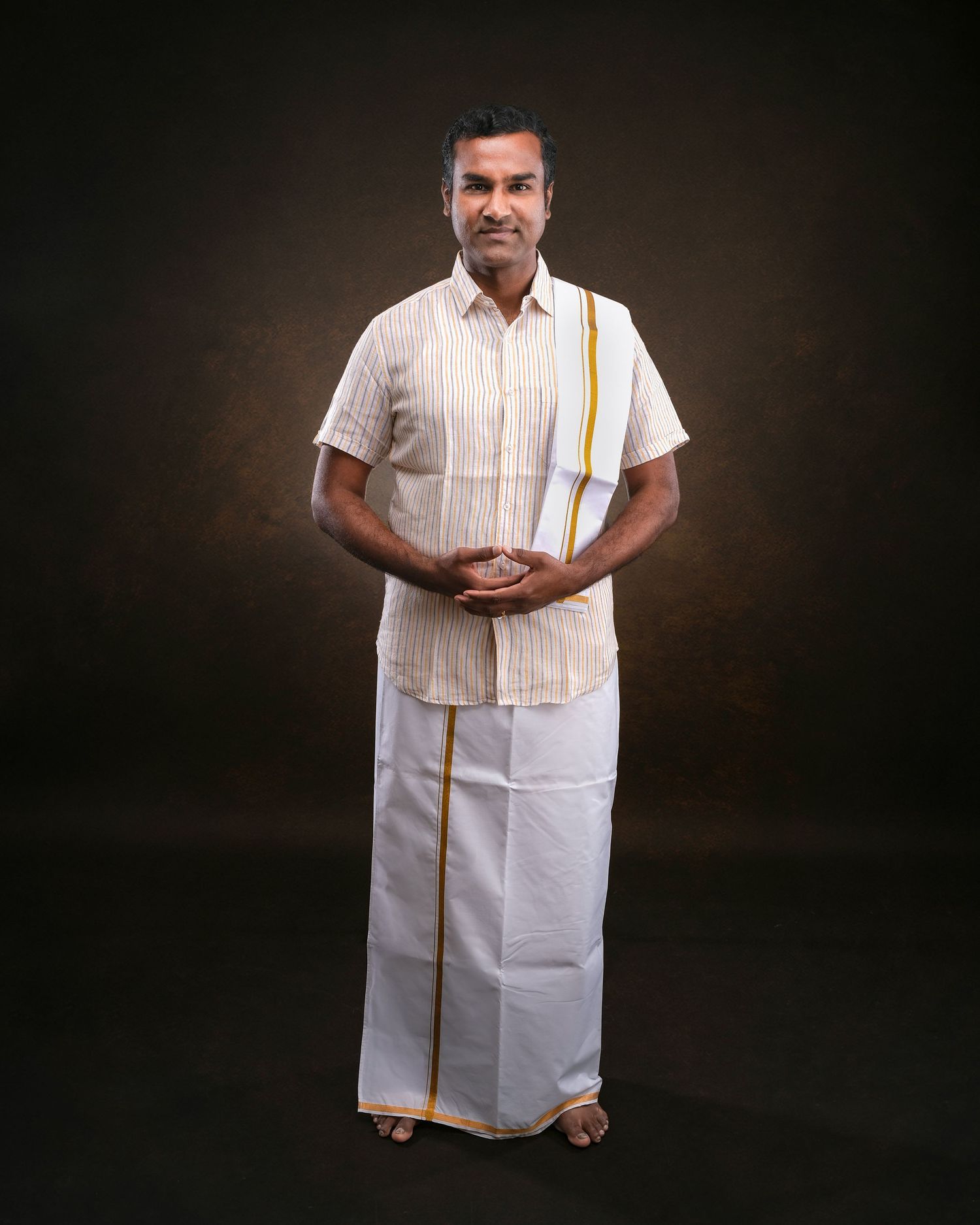 Man in traditional South Indian attire wearing white dhoti and striped shirt with yellow border standing against dark background.