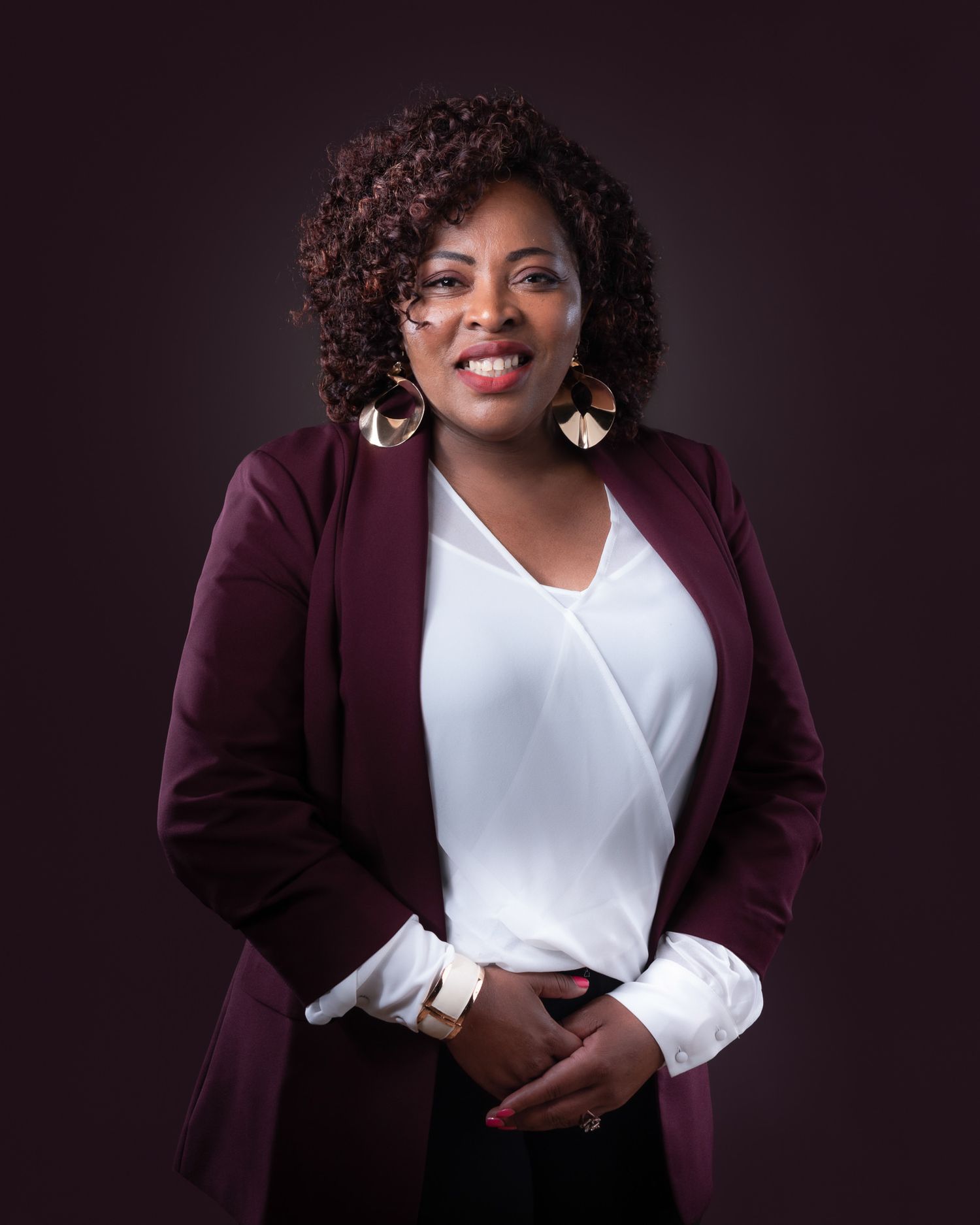 Professional portrait of someone wearing a burgundy blazer and white blouse against a dark background with statement earrings.