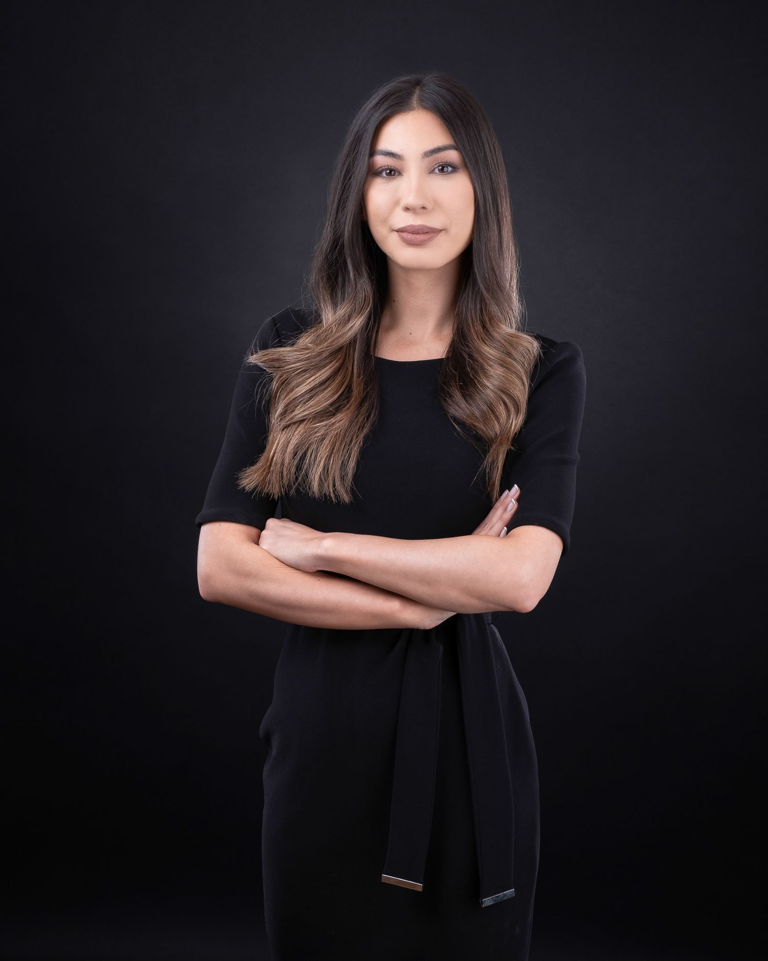Professional studio portrait of someone with long wavy brown hair wearing a black outfit against dark background, arms crossed.