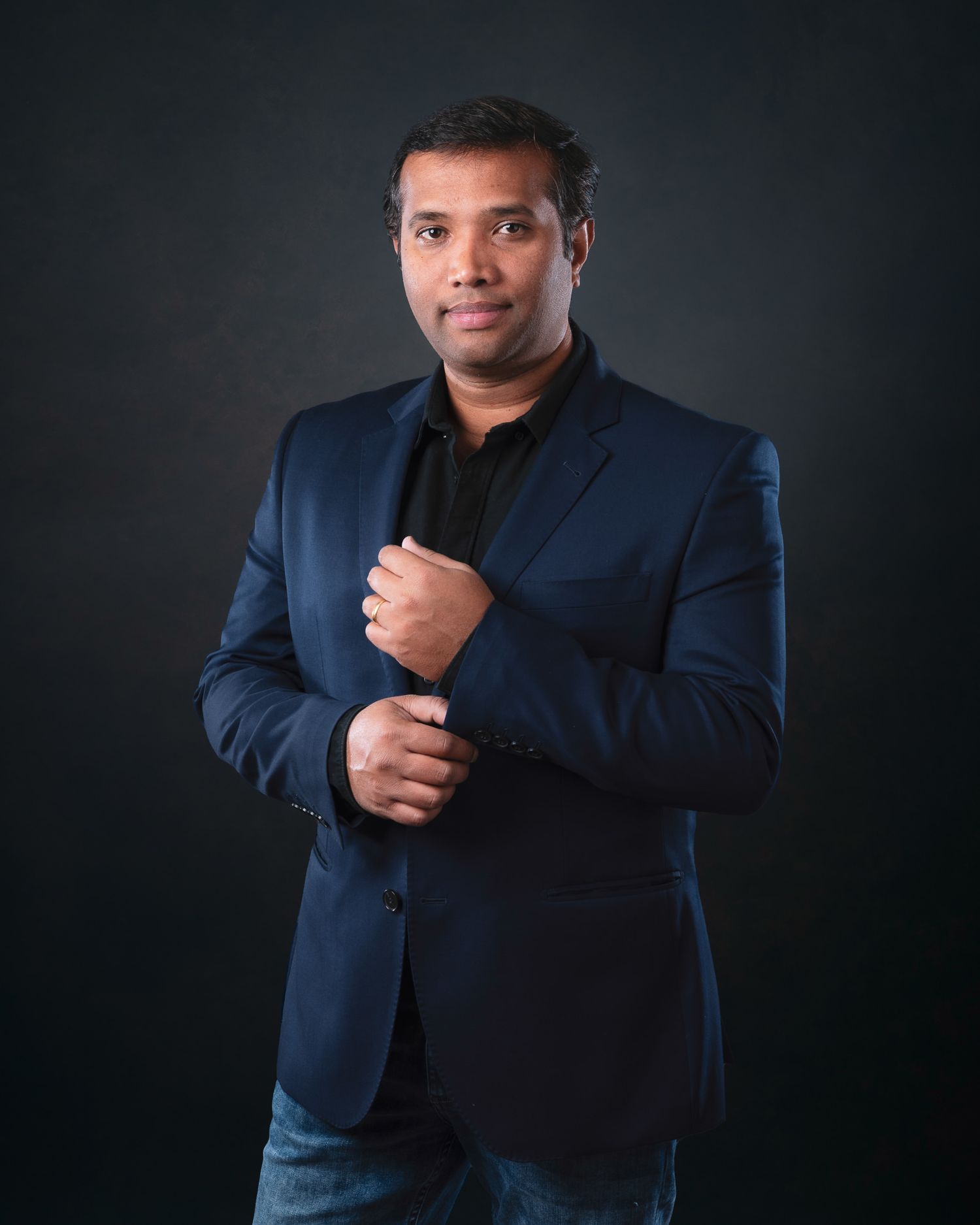 Professional headshot of a person in a navy blazer and black shirt against dark background.