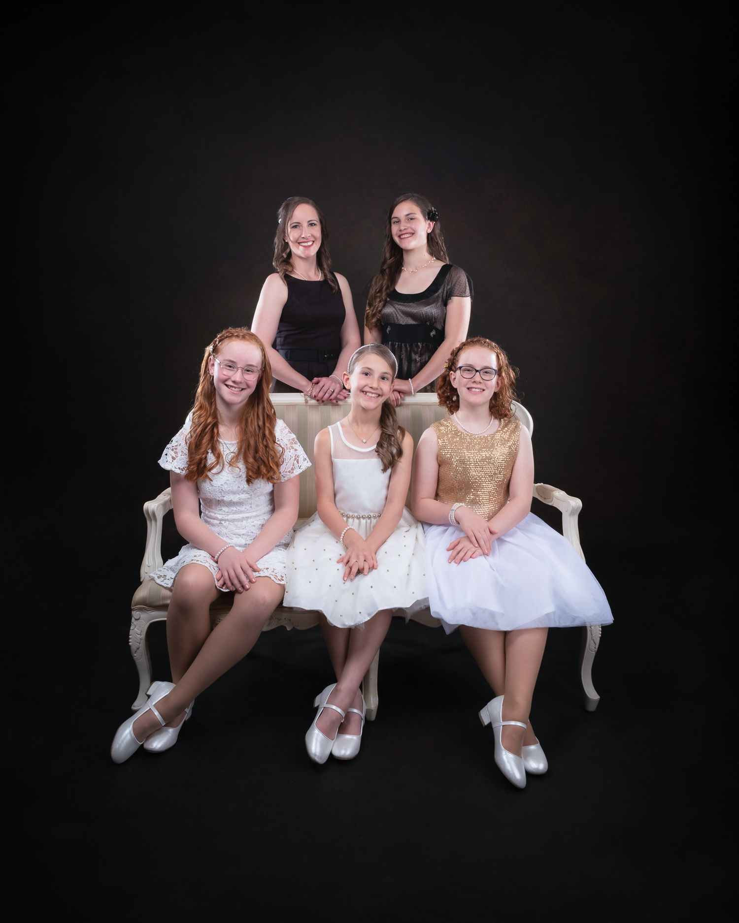 Studio portrait of dancers in ballet attire posing together on an elegant white bench against dark background.
