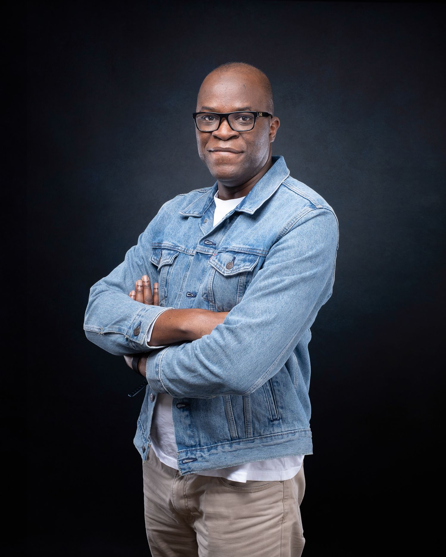 Person in denim jacket and glasses posing confidently with arms crossed against dark studio background.