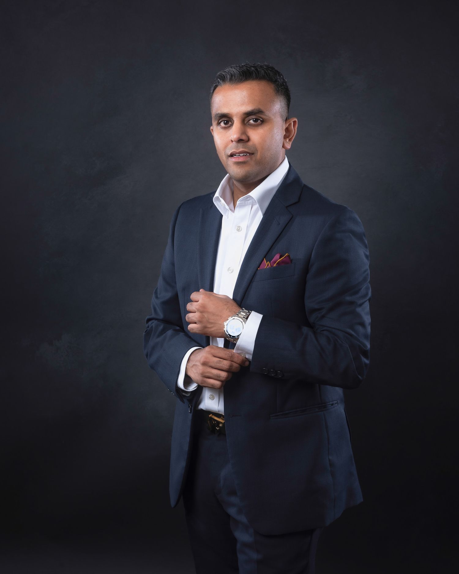 Professional portrait of a person in a navy suit and white shirt with red pocket square against dark backdrop.