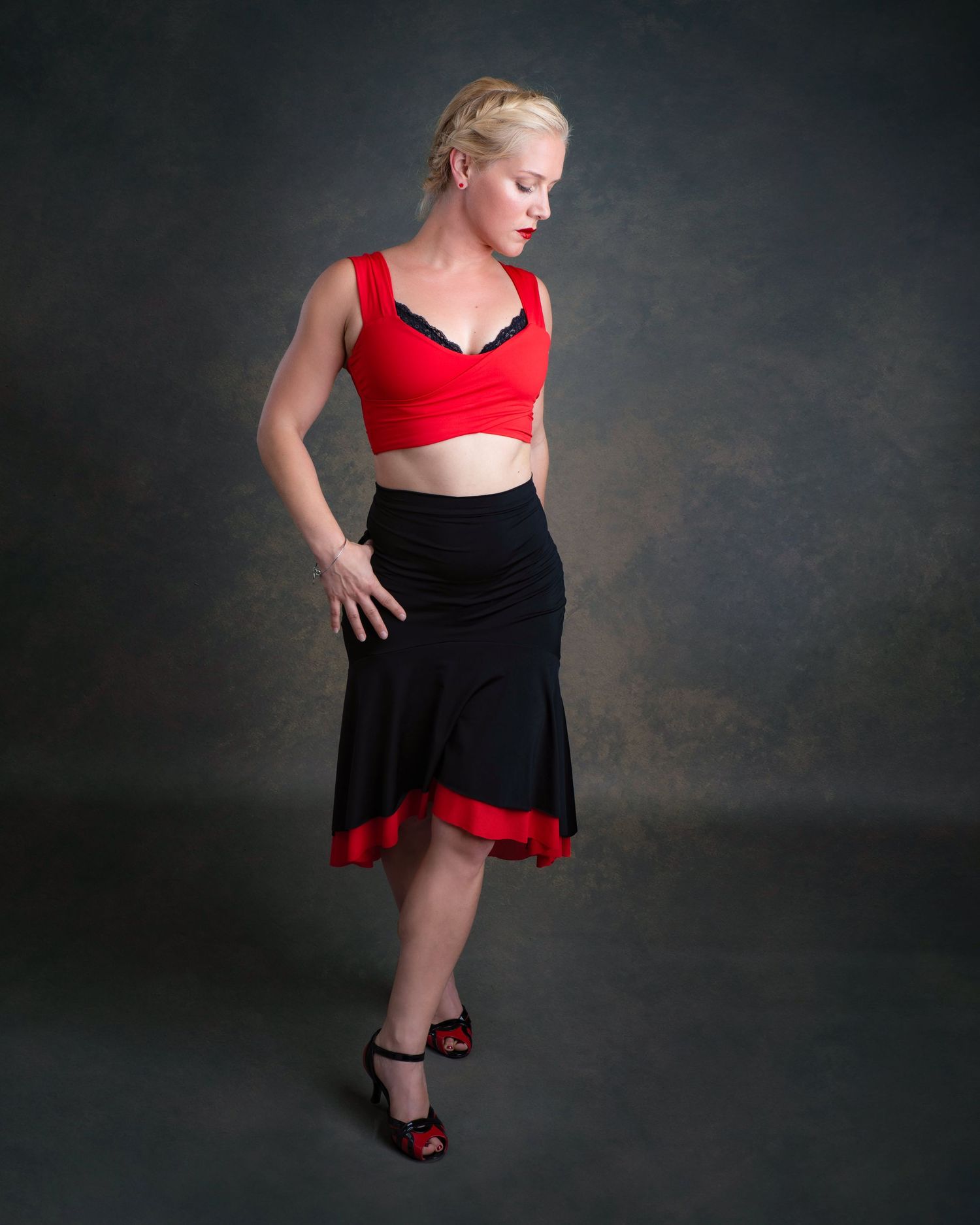A dancer in a red crop top and black skirt with red trim poses dramatically against a dark studio backdrop.