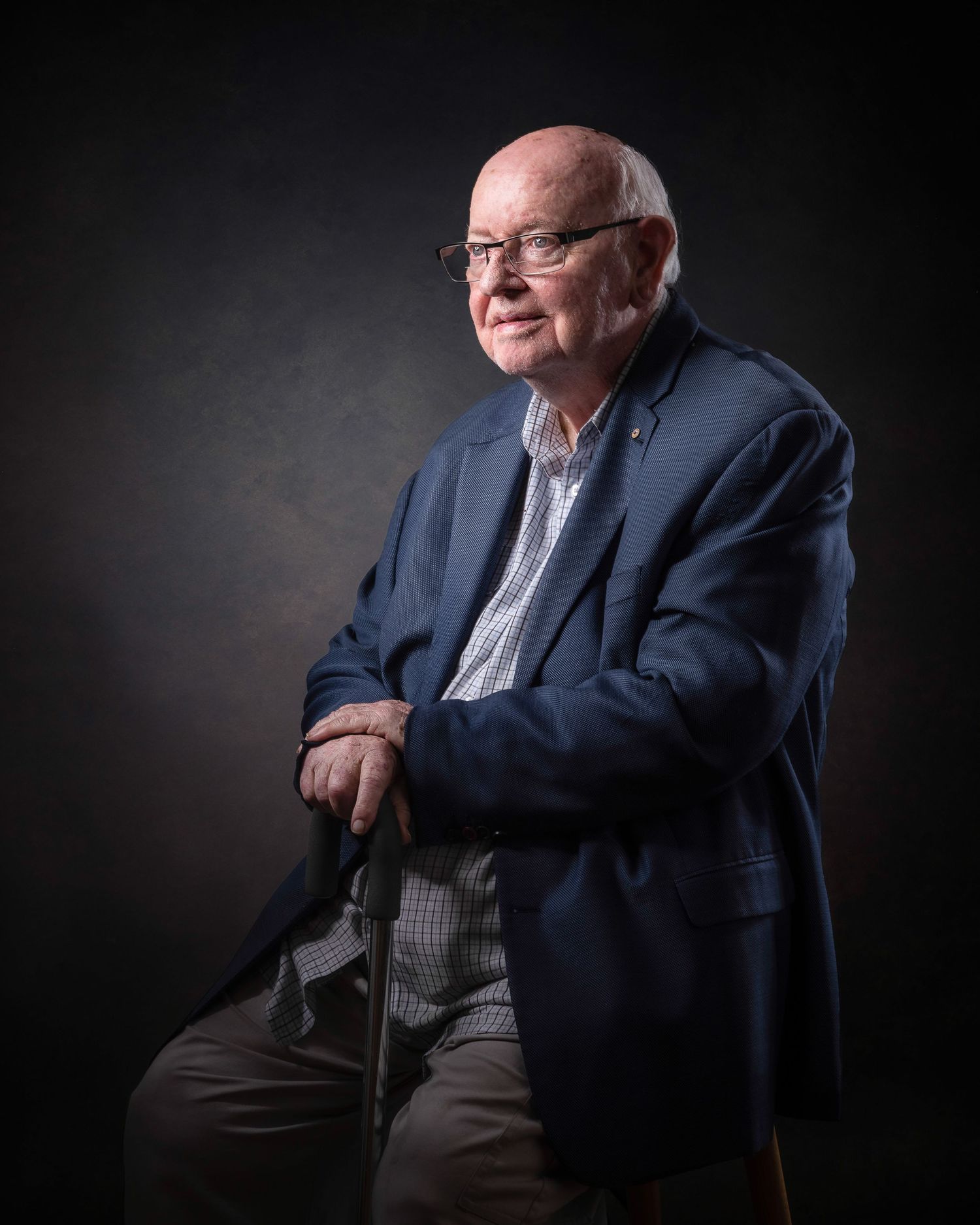 Professional studio portrait of someone in a navy blazer and glasses seated against a dark background in dramatic lighting.