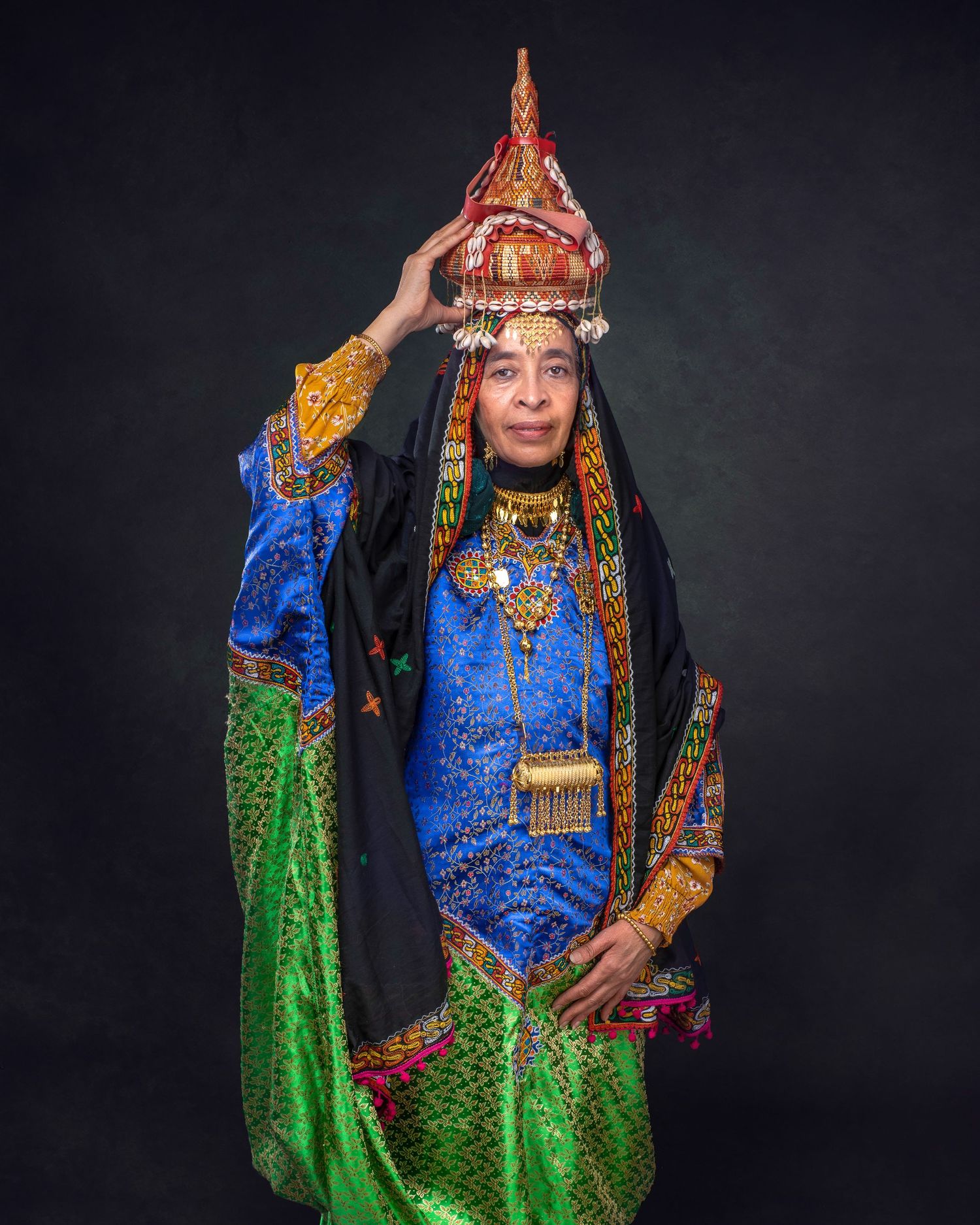 Traditional ethnic costume featuring an ornate headdress, blue and green garments with gold details against dark background.