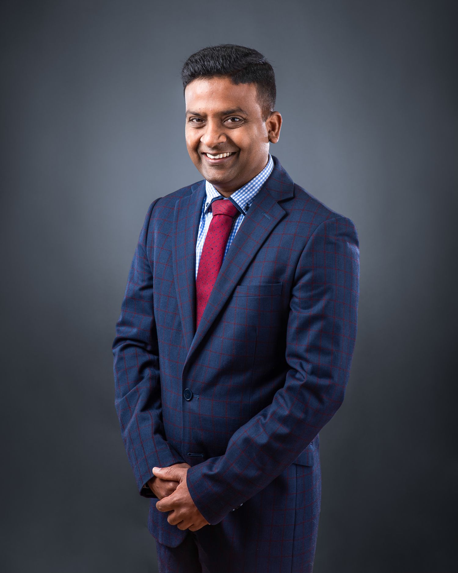 Professional studio portrait of person in navy suit with red tie against dark background.