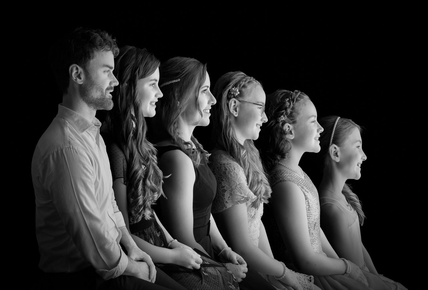 A black and white profile portrait of six people arranged in a line, looking straight ahead with dramatic lighting.