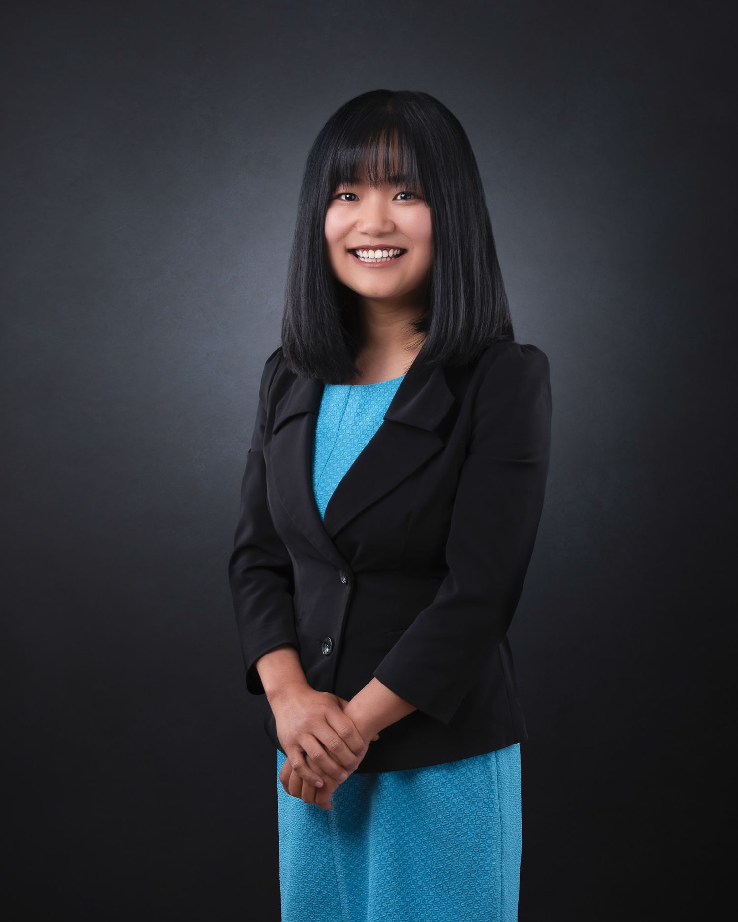 Professional studio portrait of a person wearing a black blazer and bright blue dress against a dark background.