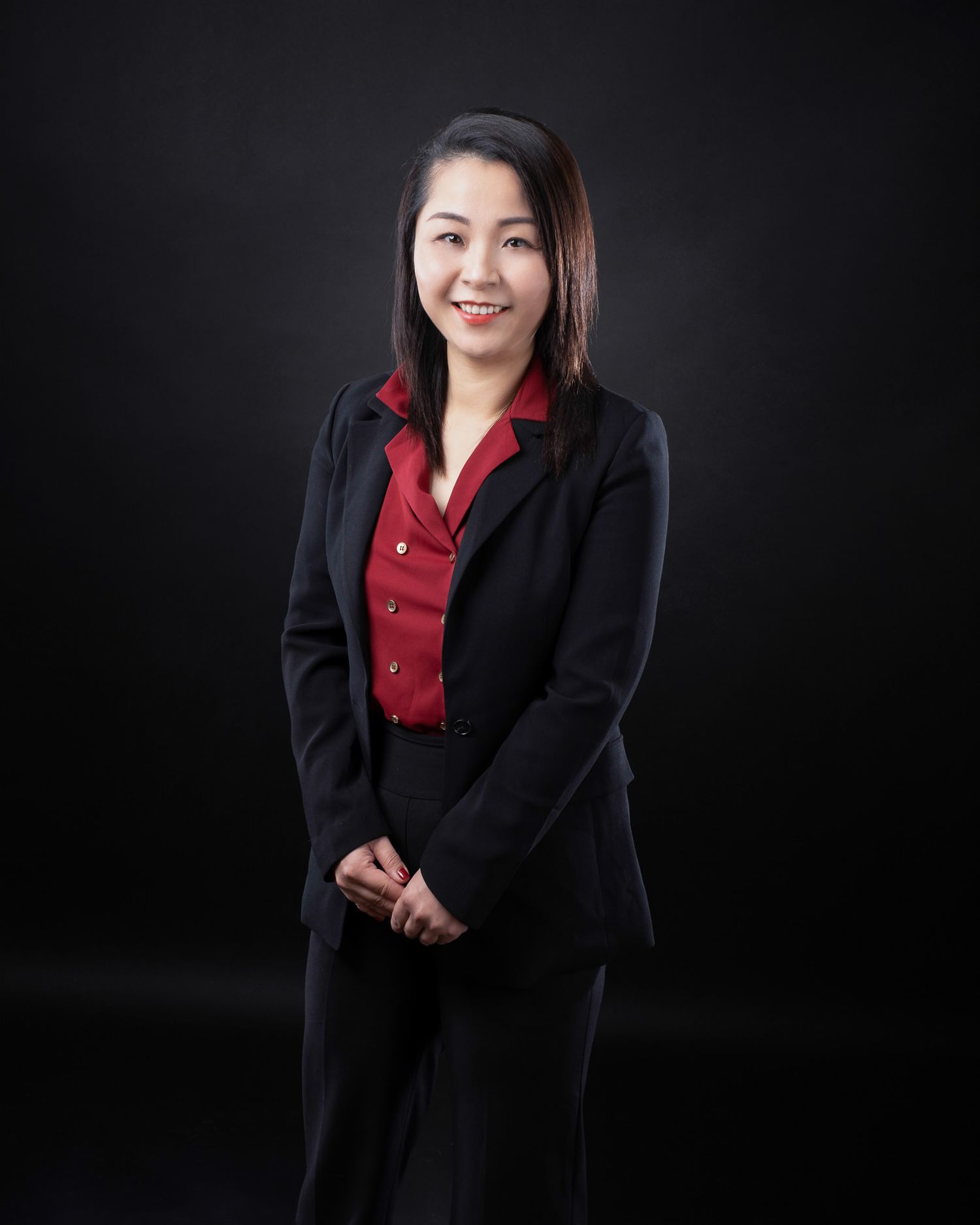 Professional corporate portrait of person wearing black blazer and red blouse against dark background.
