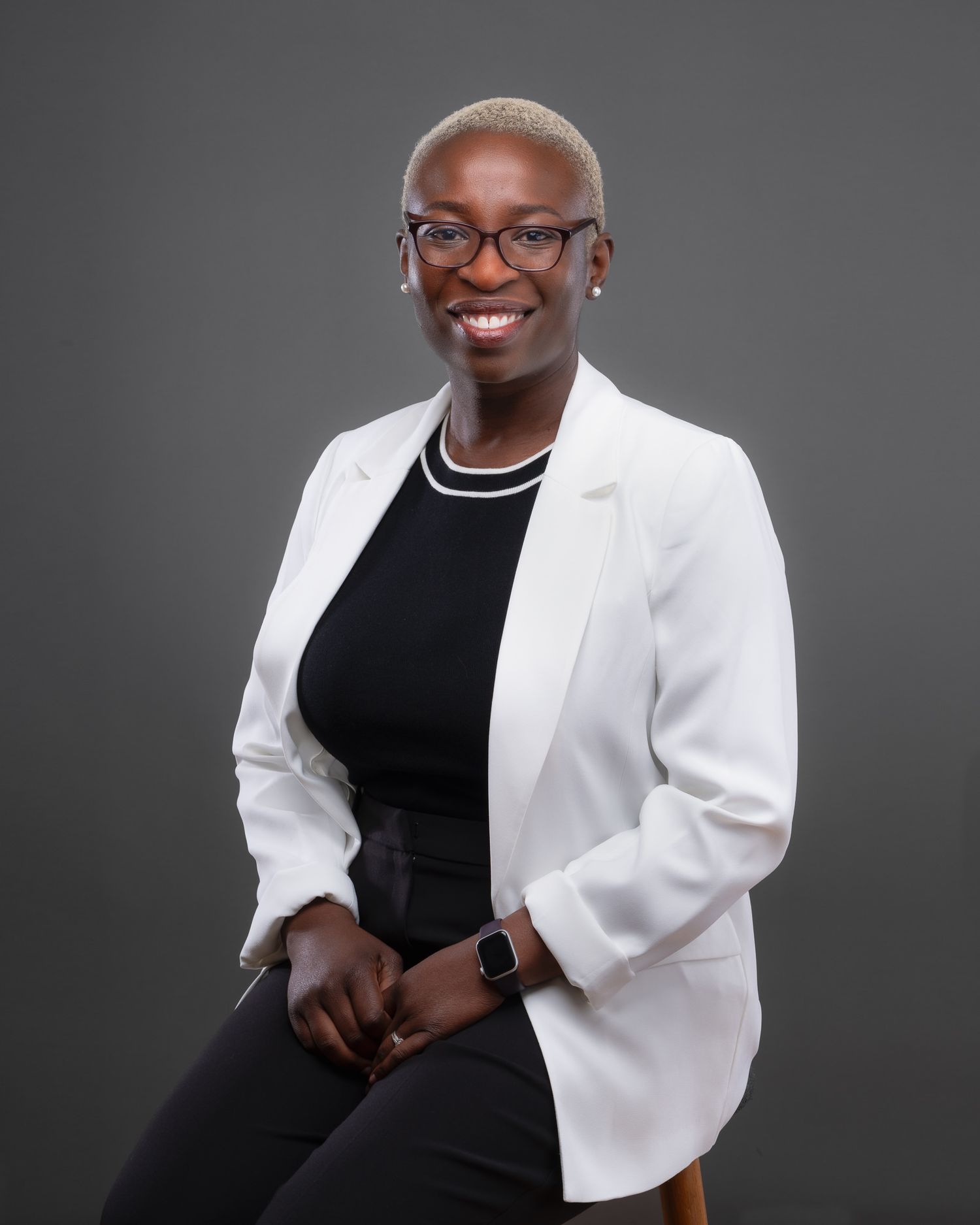 Professional studio portrait of a person with platinum blonde hair and glasses wearing a white blazer against a dark background.