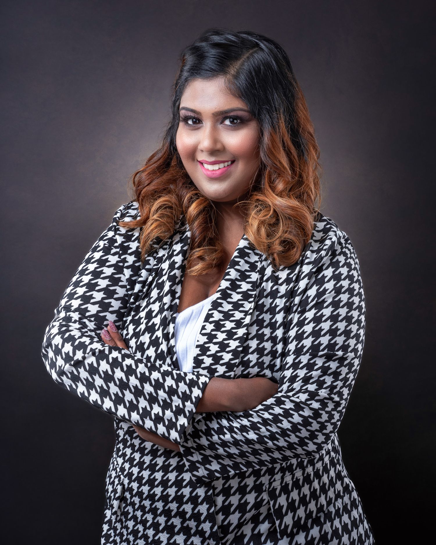 Professional portrait of person in black and white houndstooth blazer with long ombre curled hair against dark background.