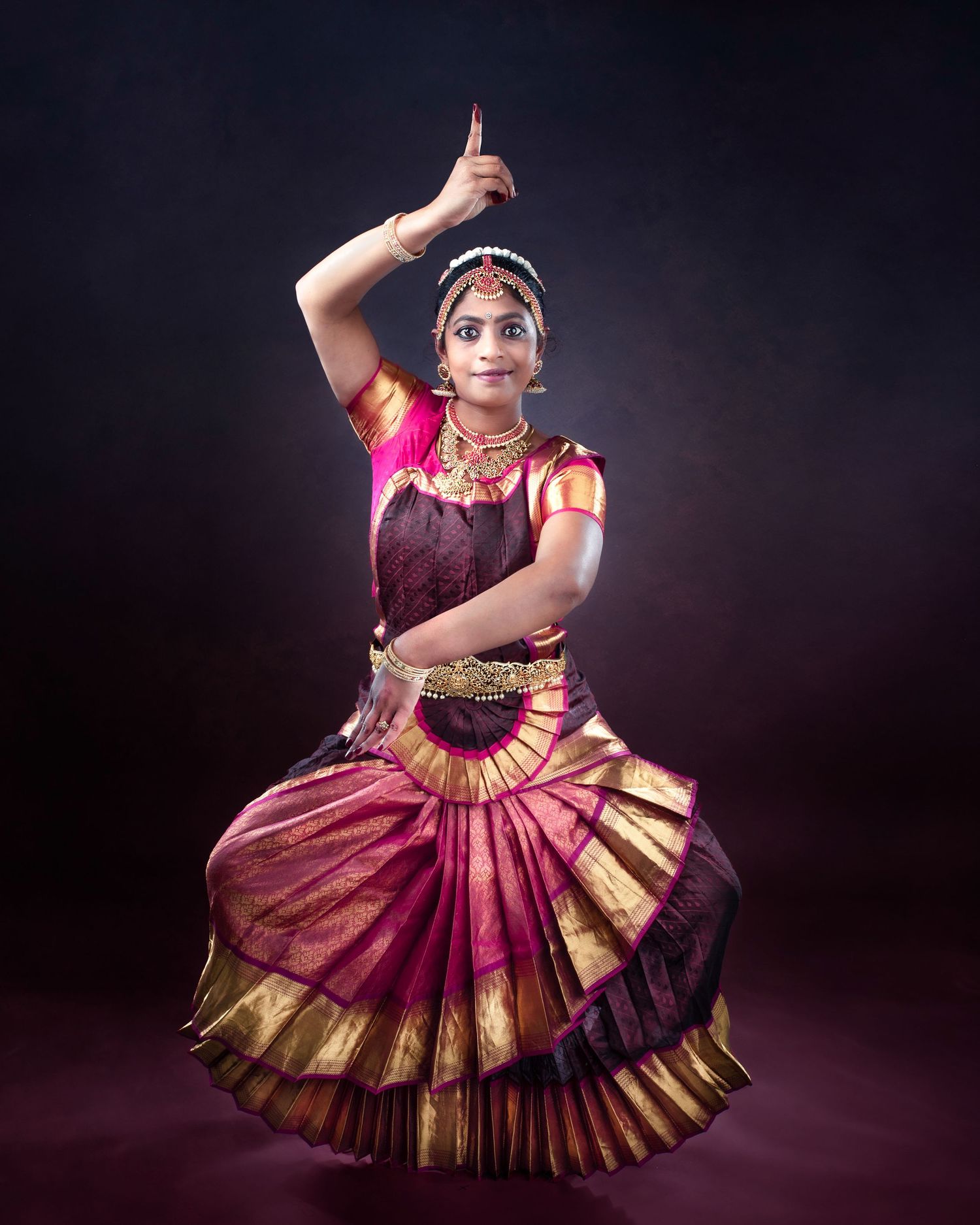 Traditional Indian classical dancer wearing ornate magenta and gold Bharatanatyam costume performs elegant pose on dark stage.