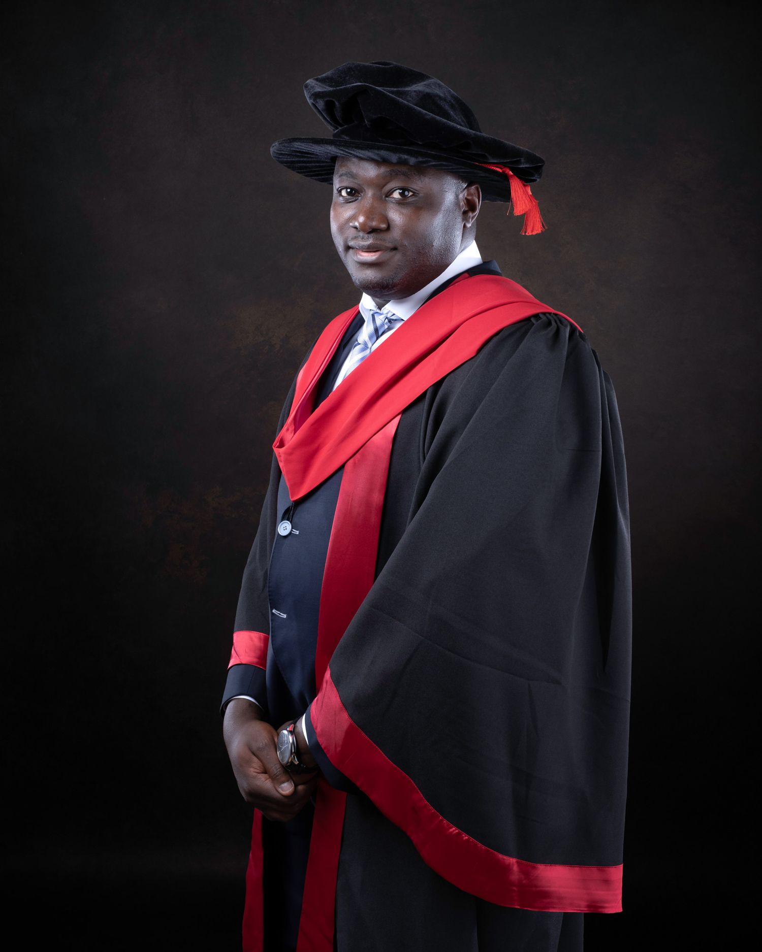 A graduate in academic regalia wearing black doctoral robes with red trim poses for a formal portrait against a dark background.