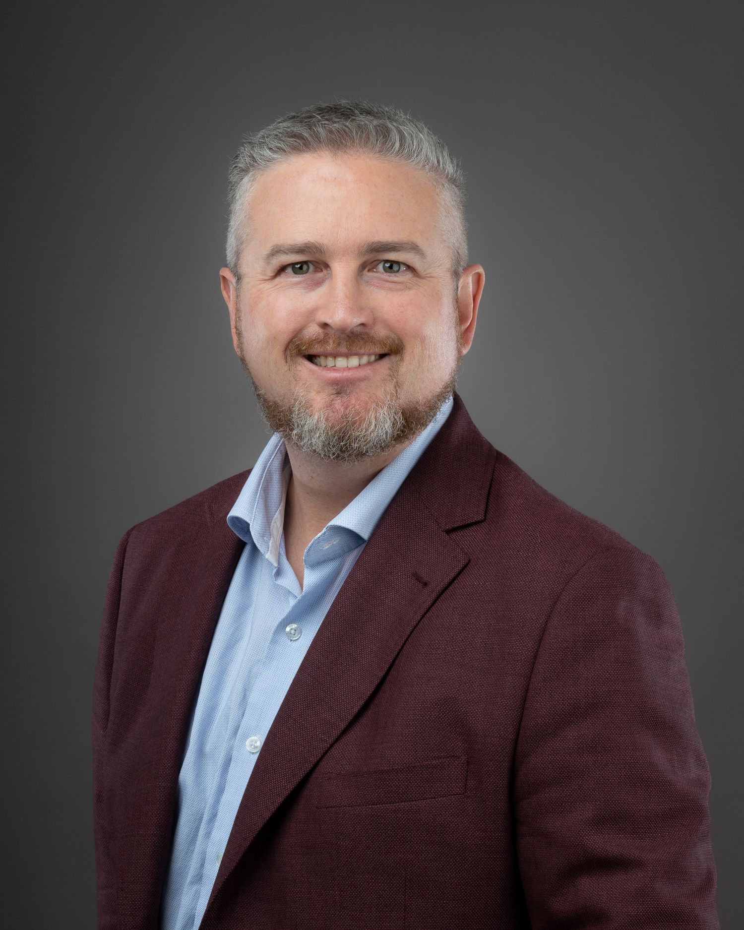 Professional headshot of person in burgundy blazer and light blue dress shirt smiling against grey background.