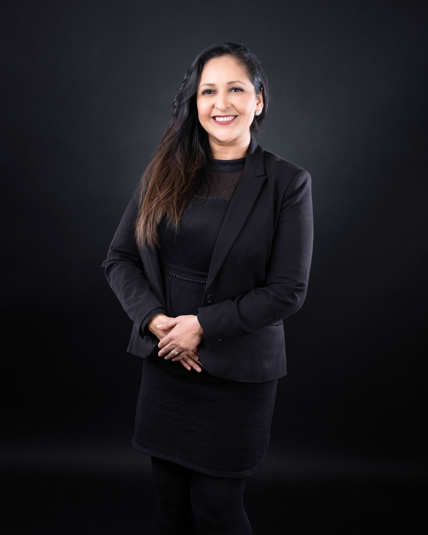Professional portrait of person in black business attire smiling warmly against dark backdrop.