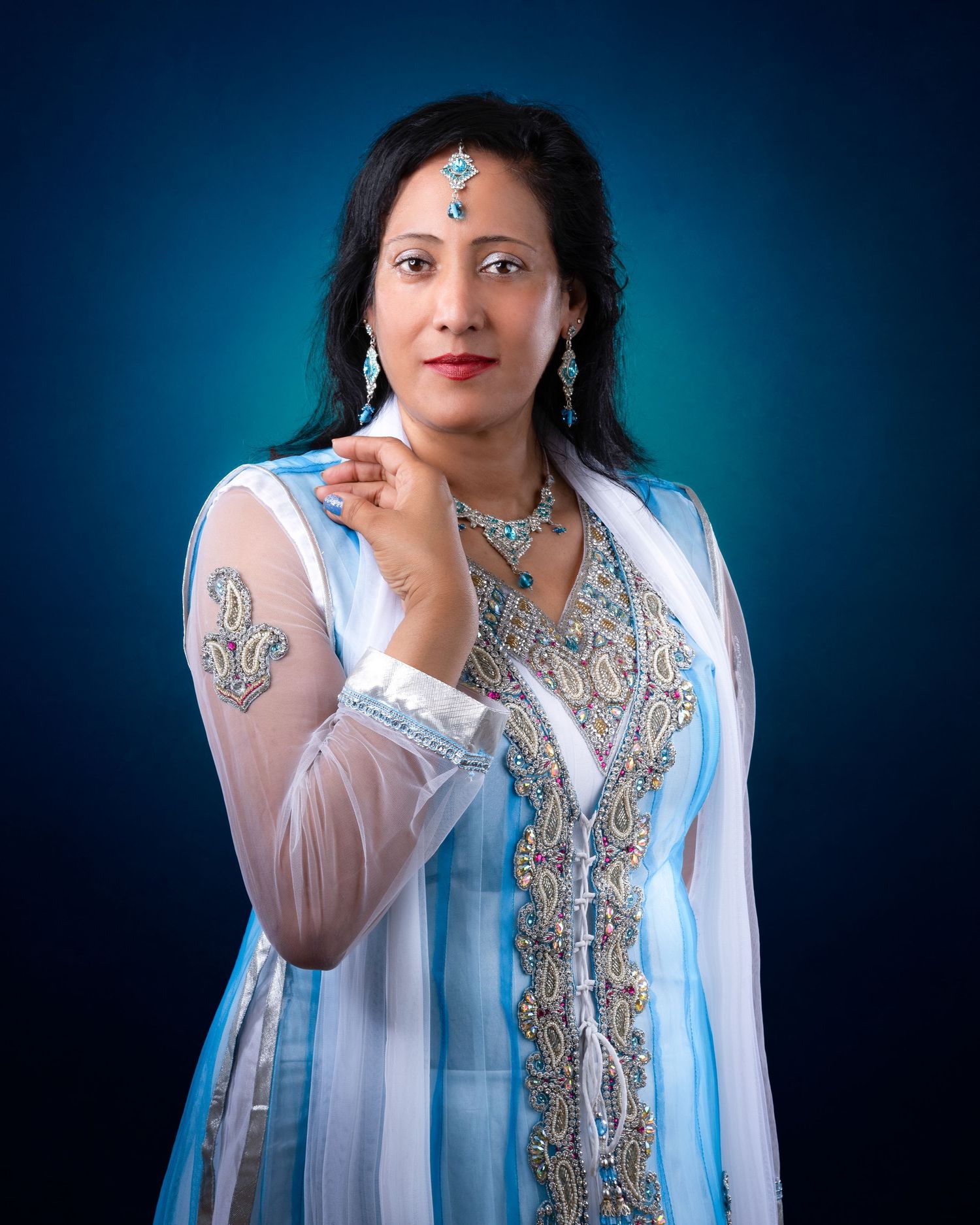 Portrait of someone wearing an embroidered white and light blue Indian outfit with silver jewelry against a blue backdrop.