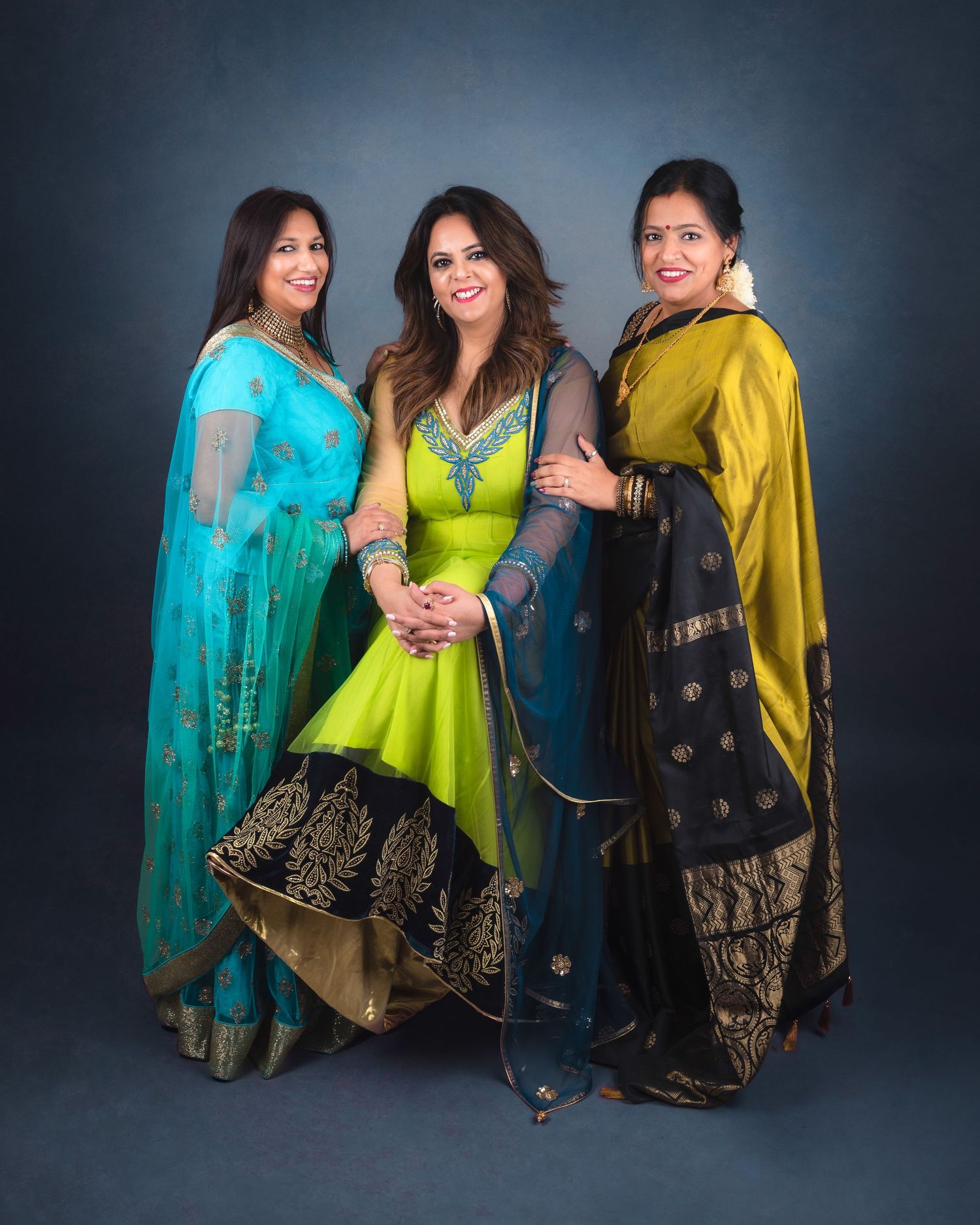 Three women in vibrant traditional Indian outfits including turquoise, lime green and gold saris pose together for a portrait.