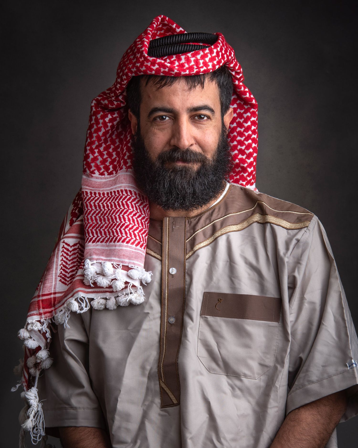 Portrait of someone wearing traditional Middle Eastern attire with red and white keffiyeh and brown thobe against dark background.