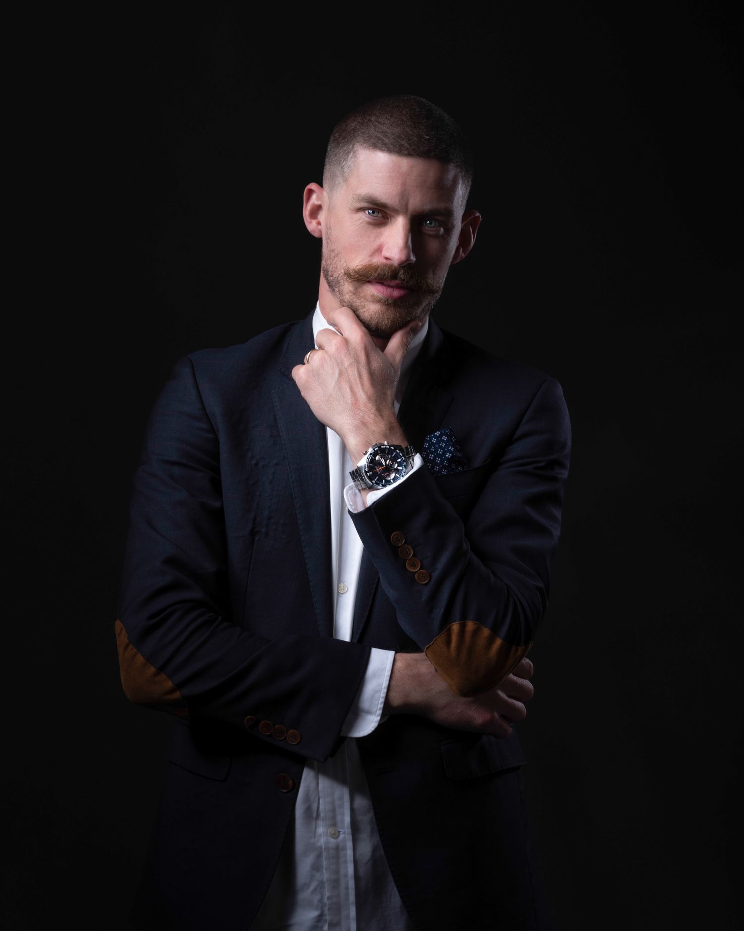 Professional portrait of person in dark suit with hand thoughtfully touching chin against black background.