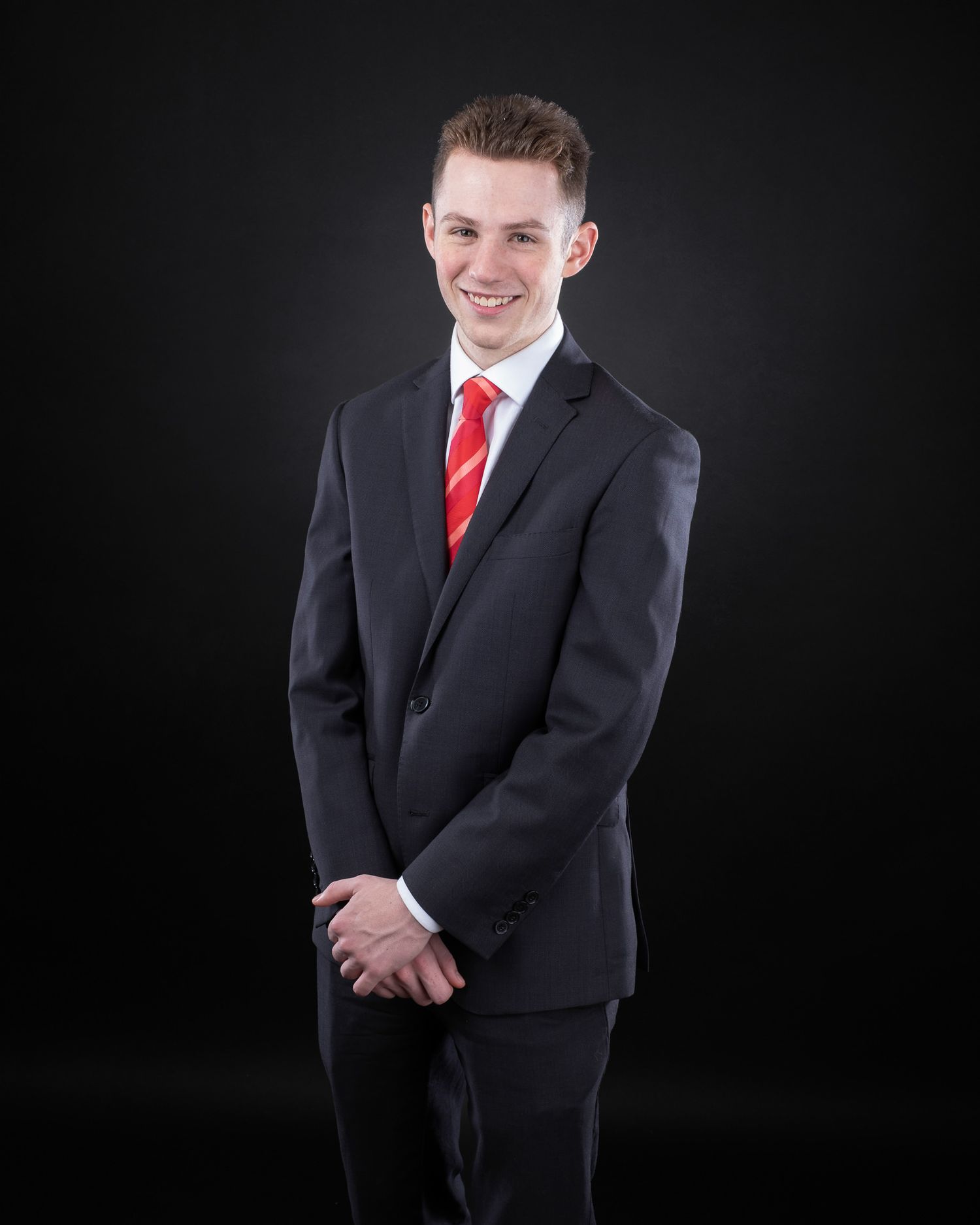 Professional portrait of person in dark suit with red tie smiling against black background.