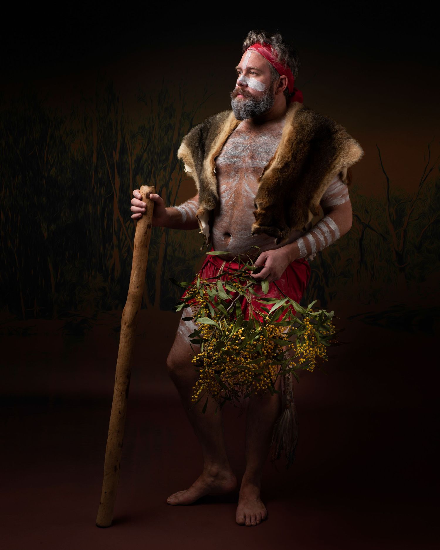 Bearded warrior in tribal attire with fur garment and walking staff stands against dark background holding greenery.