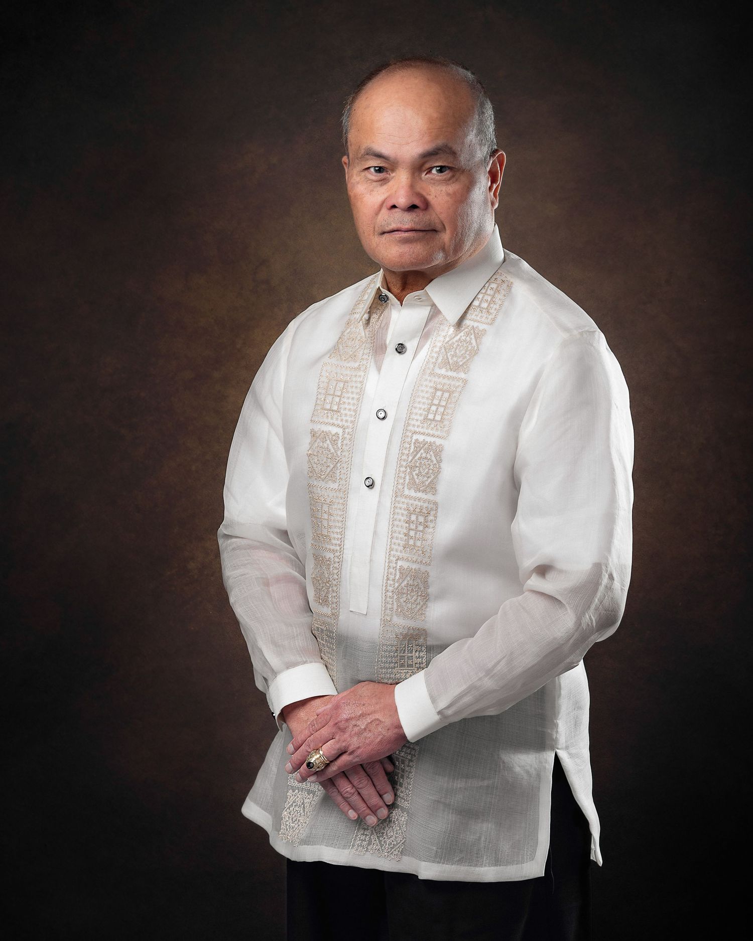 Professional portrait showing a person wearing a traditional white barong tagalog with embroidered details against a brown backdrop.
