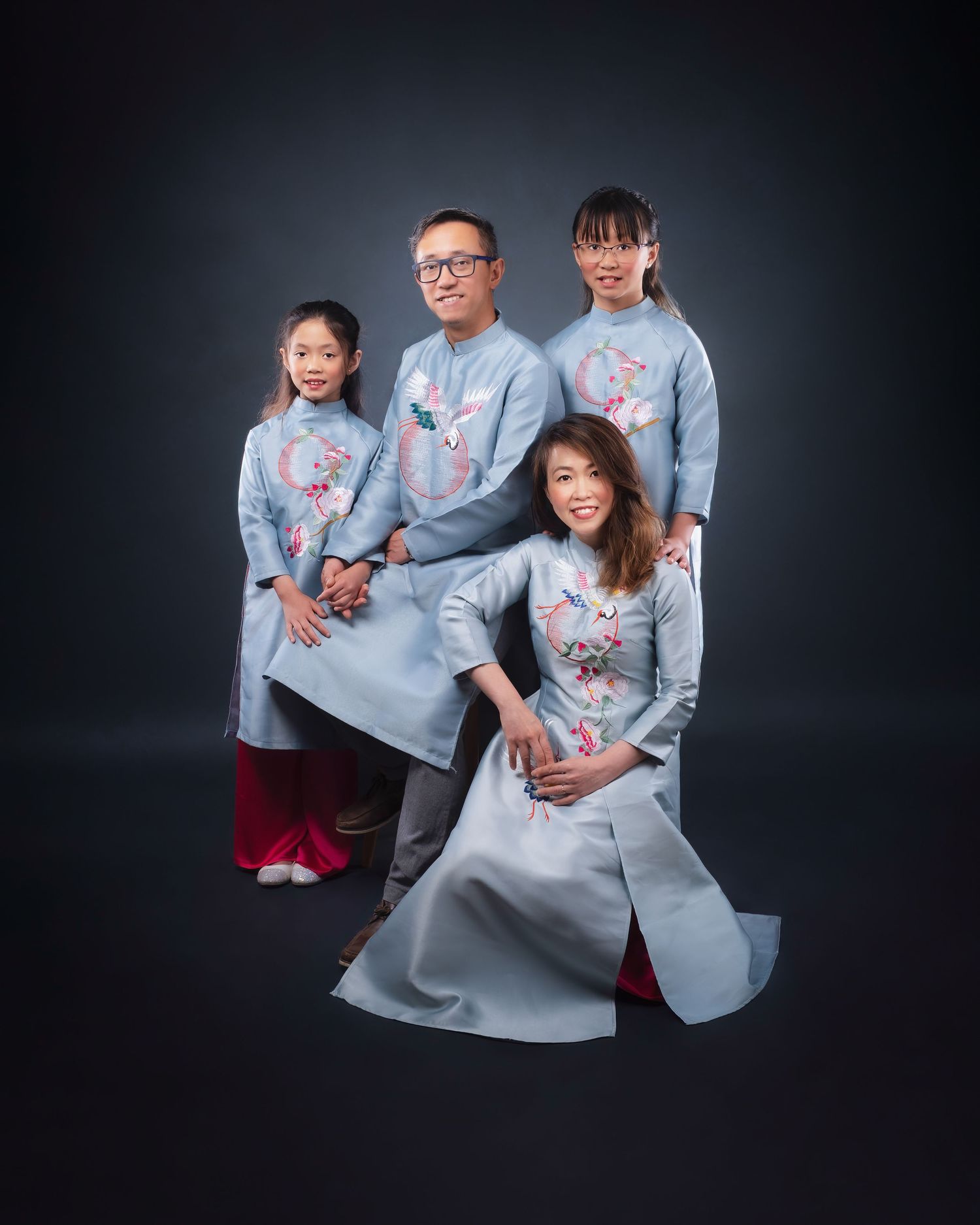 A family wearing matching light blue traditional Asian outfits with floral embroidery poses together against a dark backdrop.