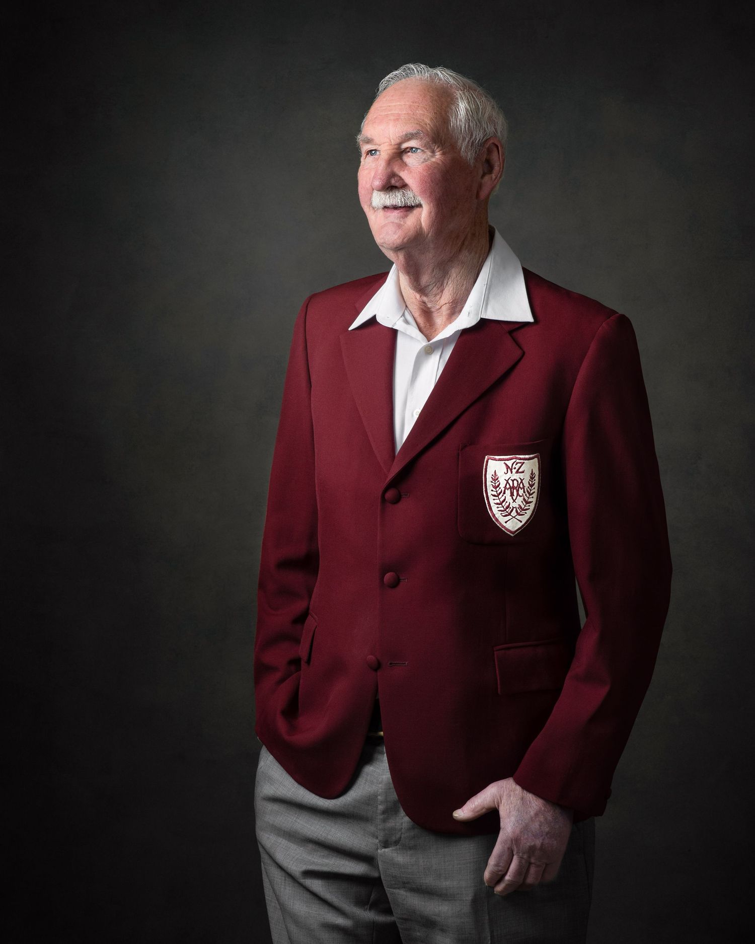 Senior figure wearing burgundy blazer with crest and white shirt poses against dark background for formal portrait.