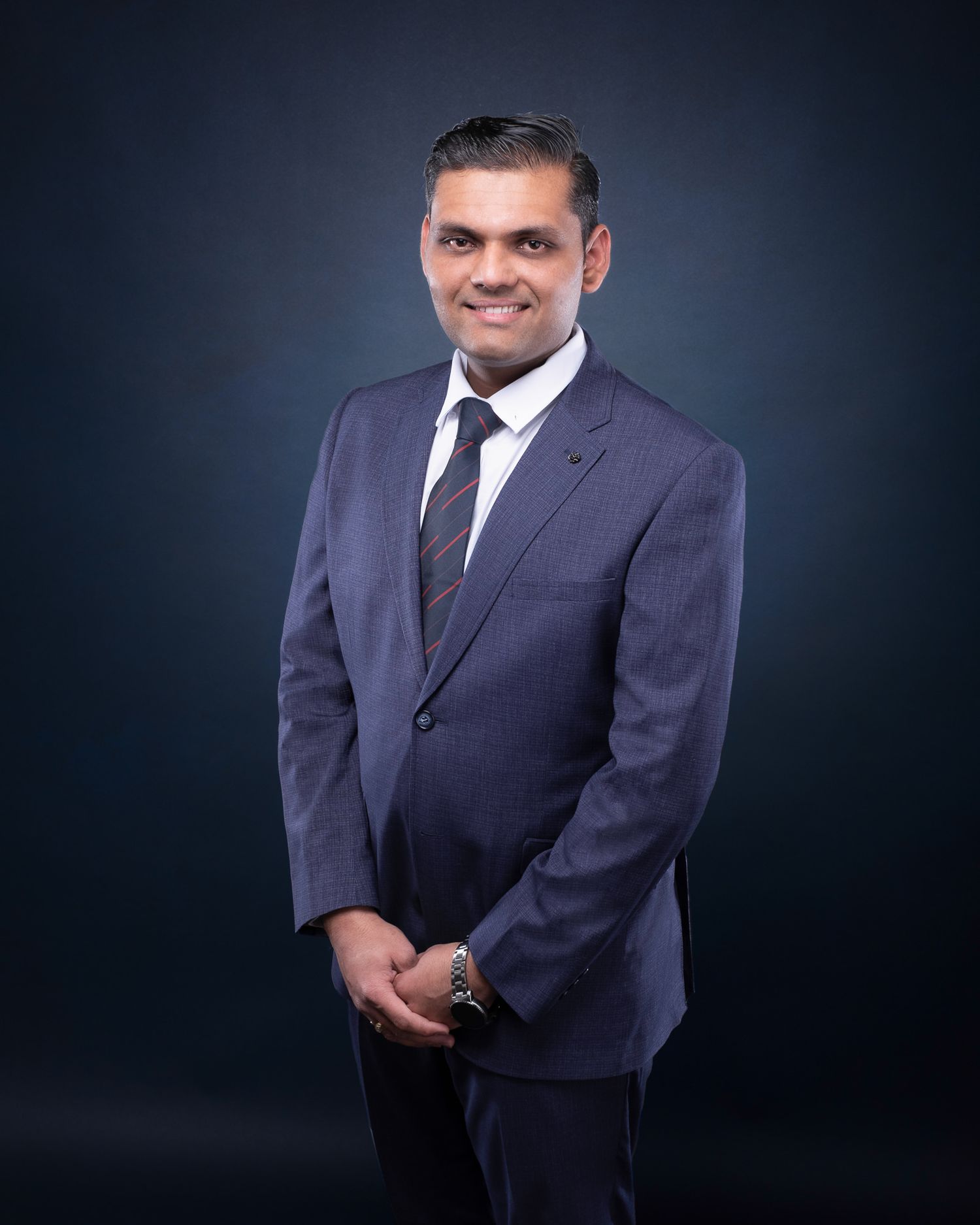 Professional portrait of a businessman in a navy suit and black tie standing against a dark background.