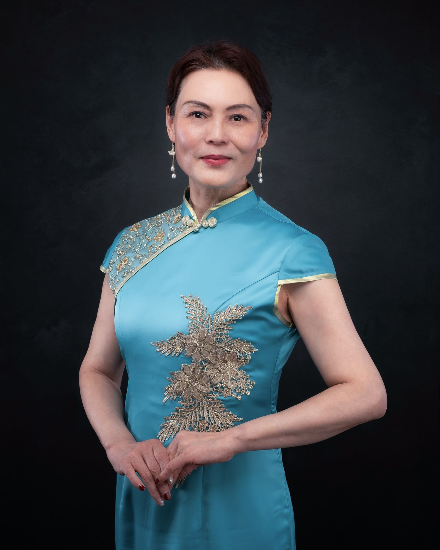 A person in a turquoise qipao dress with gold embroidered details poses against a dark background.