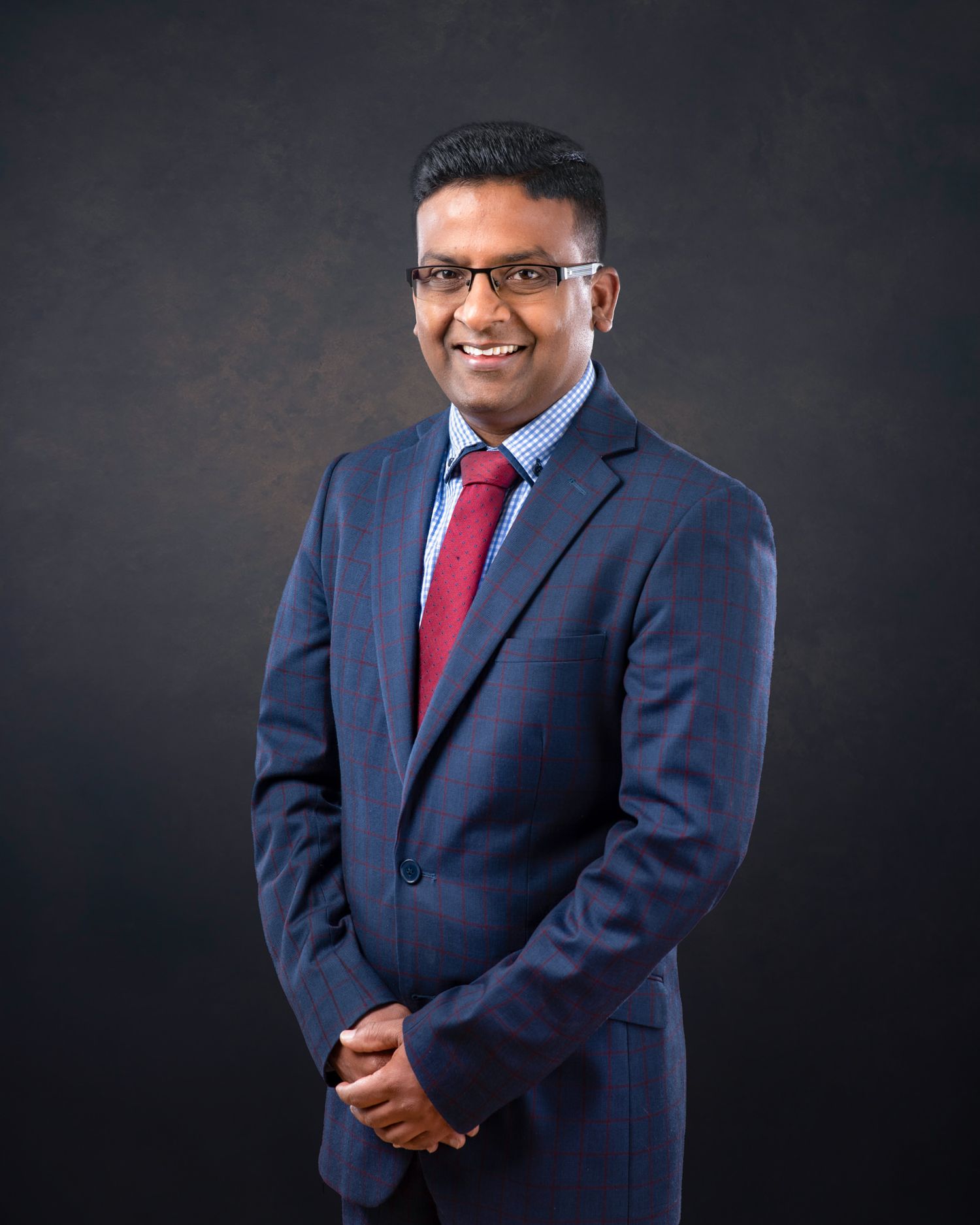 Professional portrait of someone in a navy suit and red tie standing against a dark background wearing glasses.