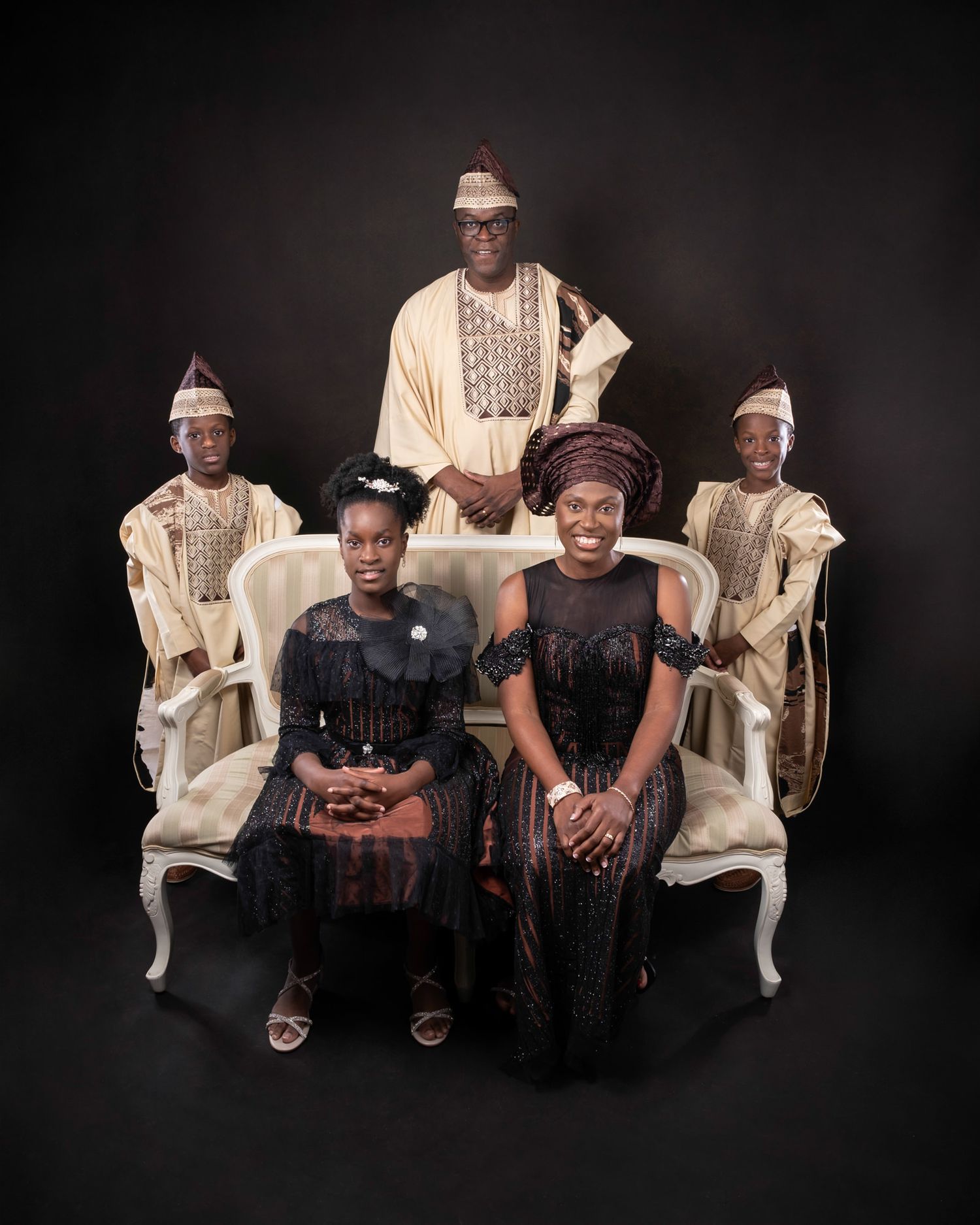 Studio portrait capturing a traditional African family posing on elegant furniture against a black background.