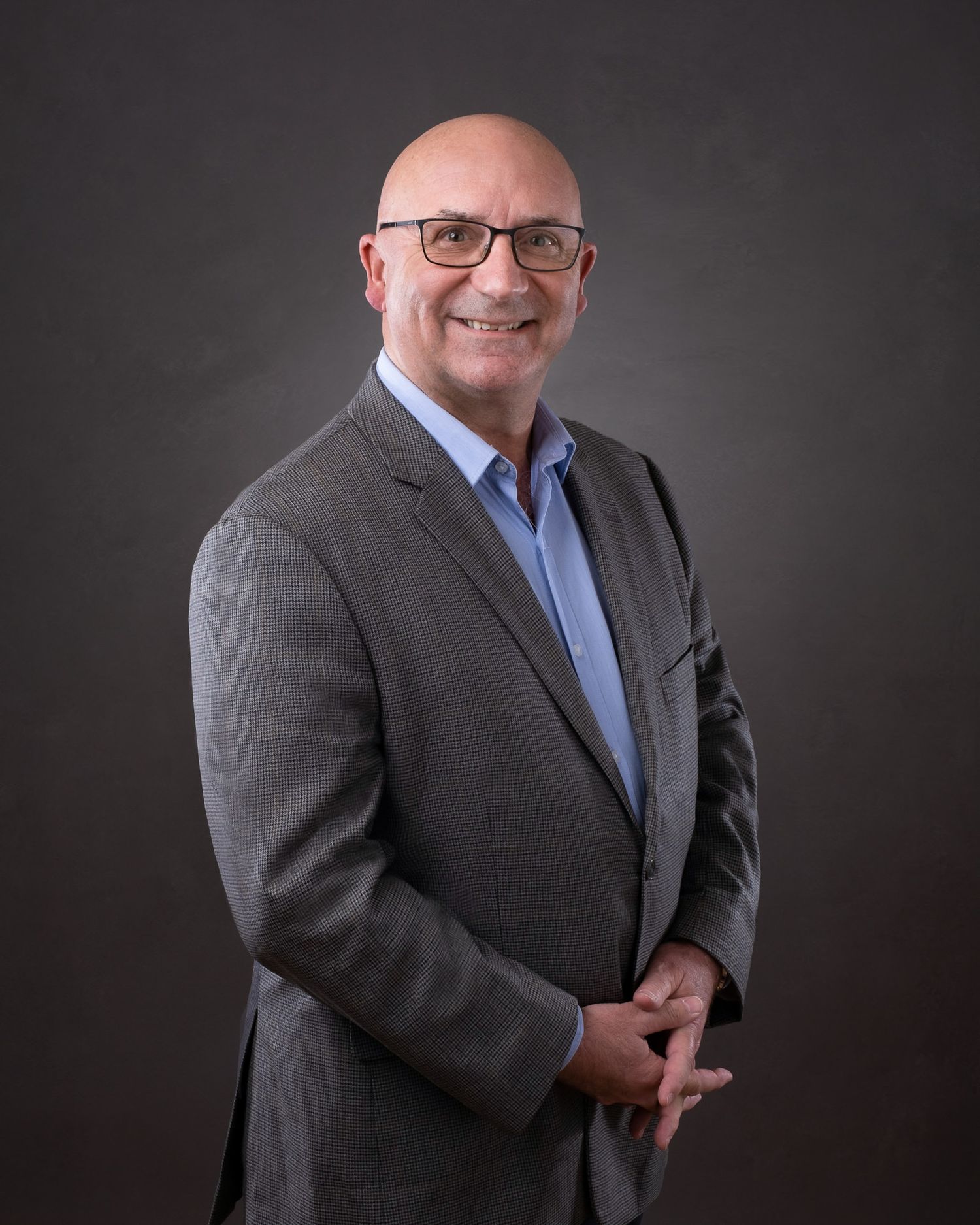 Professional studio portrait of a person in glasses and gray suit jacket against dark background with soft lighting.