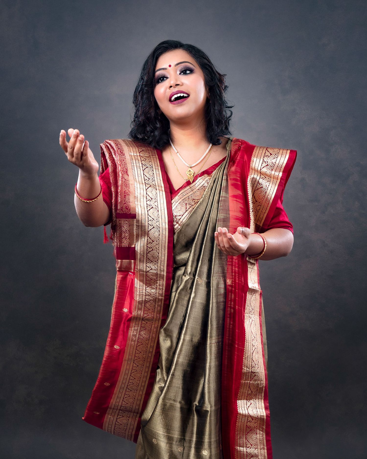 Person wearing an elegant gold and red traditional Indian saree poses expressively against a dark background.