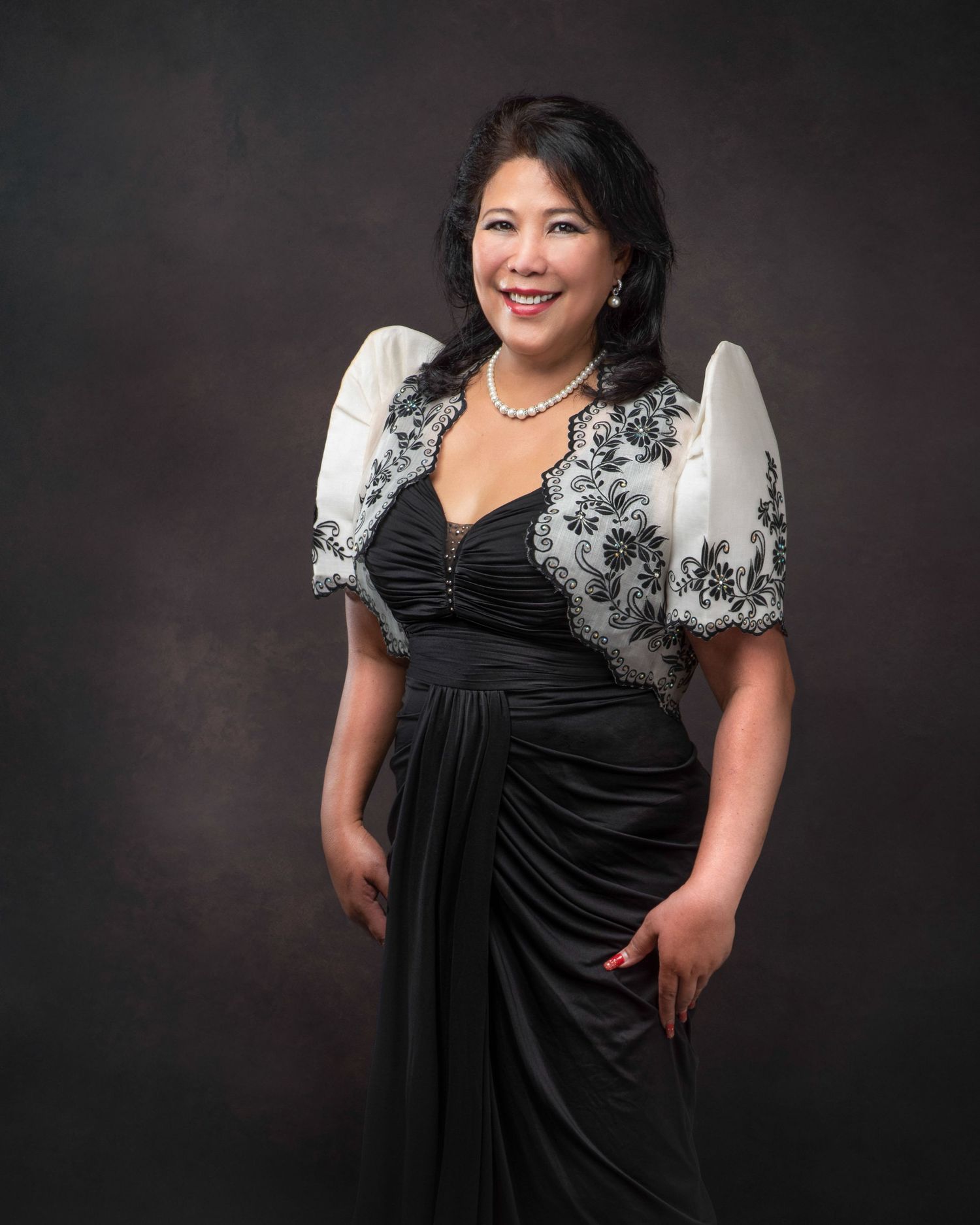 Elegant formal portrait of a person wearing a black draped evening gown with white lace bolero jacket against dark background.