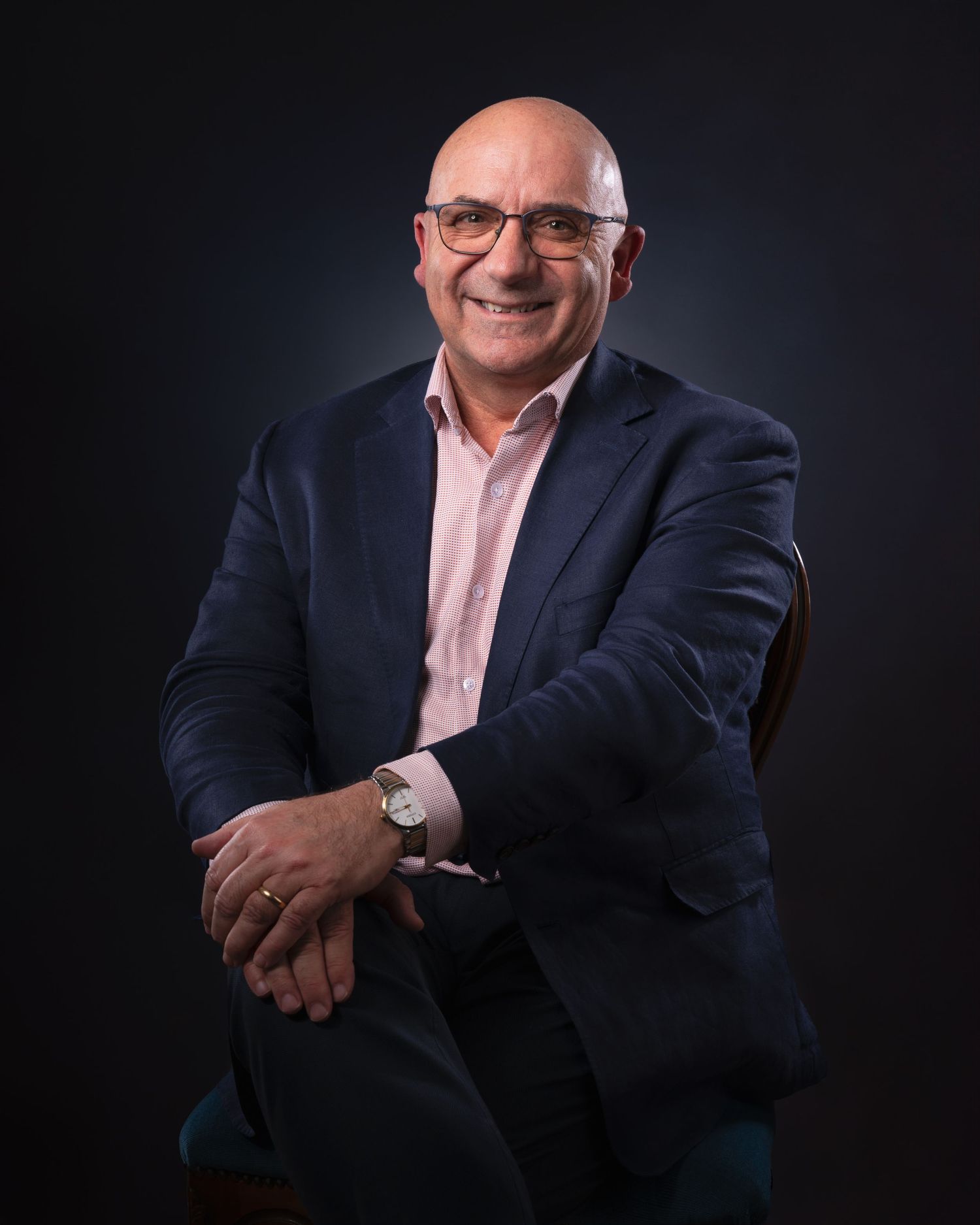 Professional studio portrait of a person wearing glasses and a pink dress shirt with navy blazer against dark background.