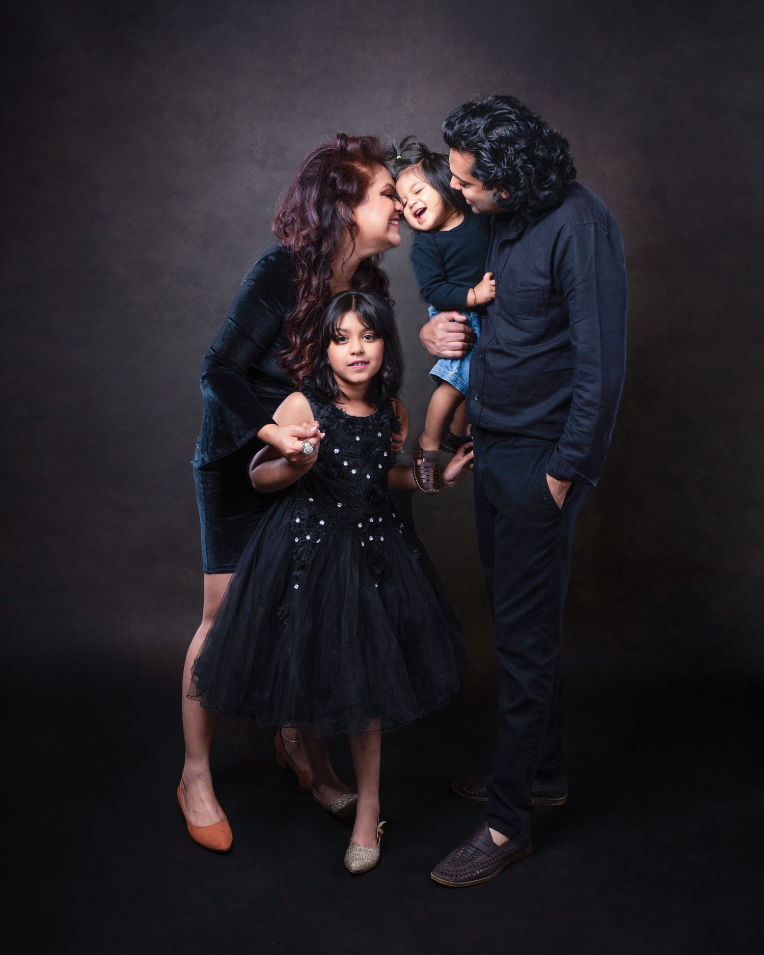 A family dressed in dark clothing poses together against a dark background for a moody studio portrait.